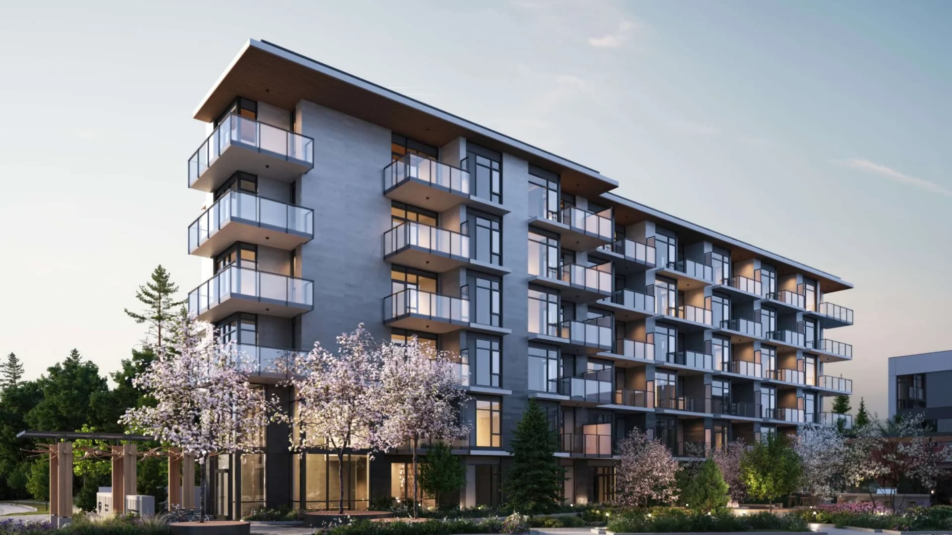 Edge Way at leləm̓ – A low-angle exterior architectural render of a modern, multi-story mid-rise residential building featuring concrete cladding, dark window frames, glass-railed balconies, and warm wood accents under the roof overhang. The landscaped ground level includes blossoming cherry trees, evergreens, subtle pathway lighting, and a timber pergola at twilight.