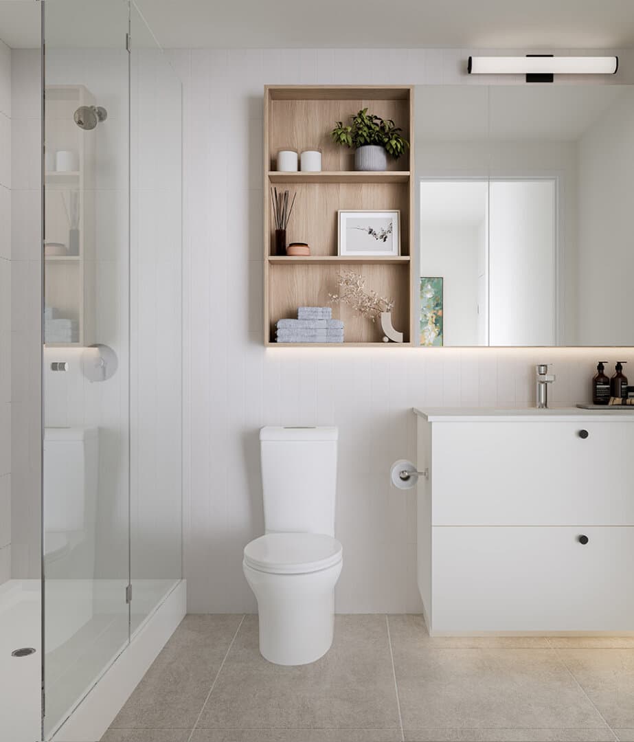 Inlet District – A bright, modern bathroom featuring a glass-enclosed walk-in shower, a white toilet, and a minimalist white vanity with black hardware. Above the toilet and counter, a light wood recessed shelving unit and a large frameless mirror with under-cabinet ambient lighting complete the clean, contemporary aesthetic.
