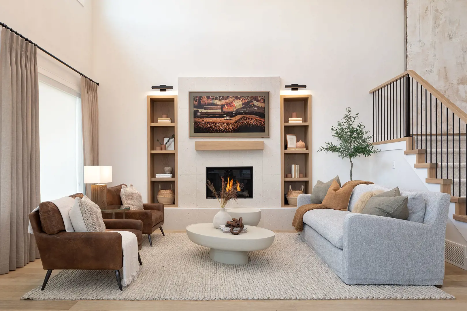 Ridgemont – Two-story living room with a tiled fireplace, built-in shelving on either side, a gray sofa and brown leather armchairs on a woven area rug, with a staircase visible on the right.