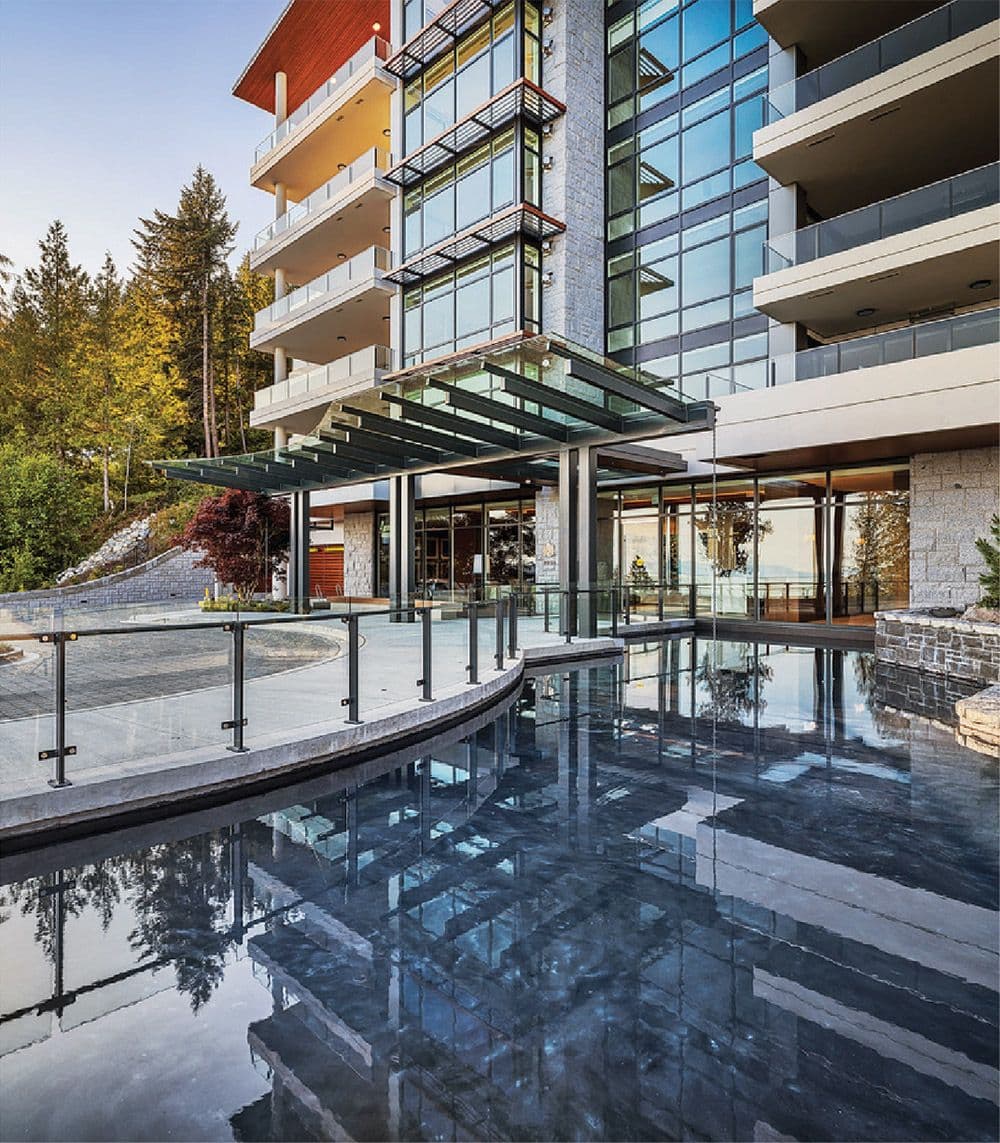 Wildwood at Uplands – Building exterior entrance with a glass canopy over a reflecting pool feature, stone accents, and a glass-walled lobby, with forested hillside behind.