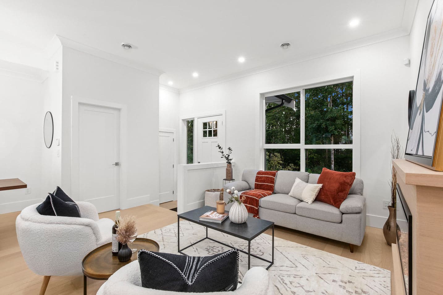 Grandview Walk – A bright, open living room staged with a grey sofa, two white textured armchairs, and a modern fireplace with a light wood mantel. Large picture windows overlook lush green trees outside, while white walls, recessed ceiling lights, and light wood flooring complete the contemporary aesthetic.