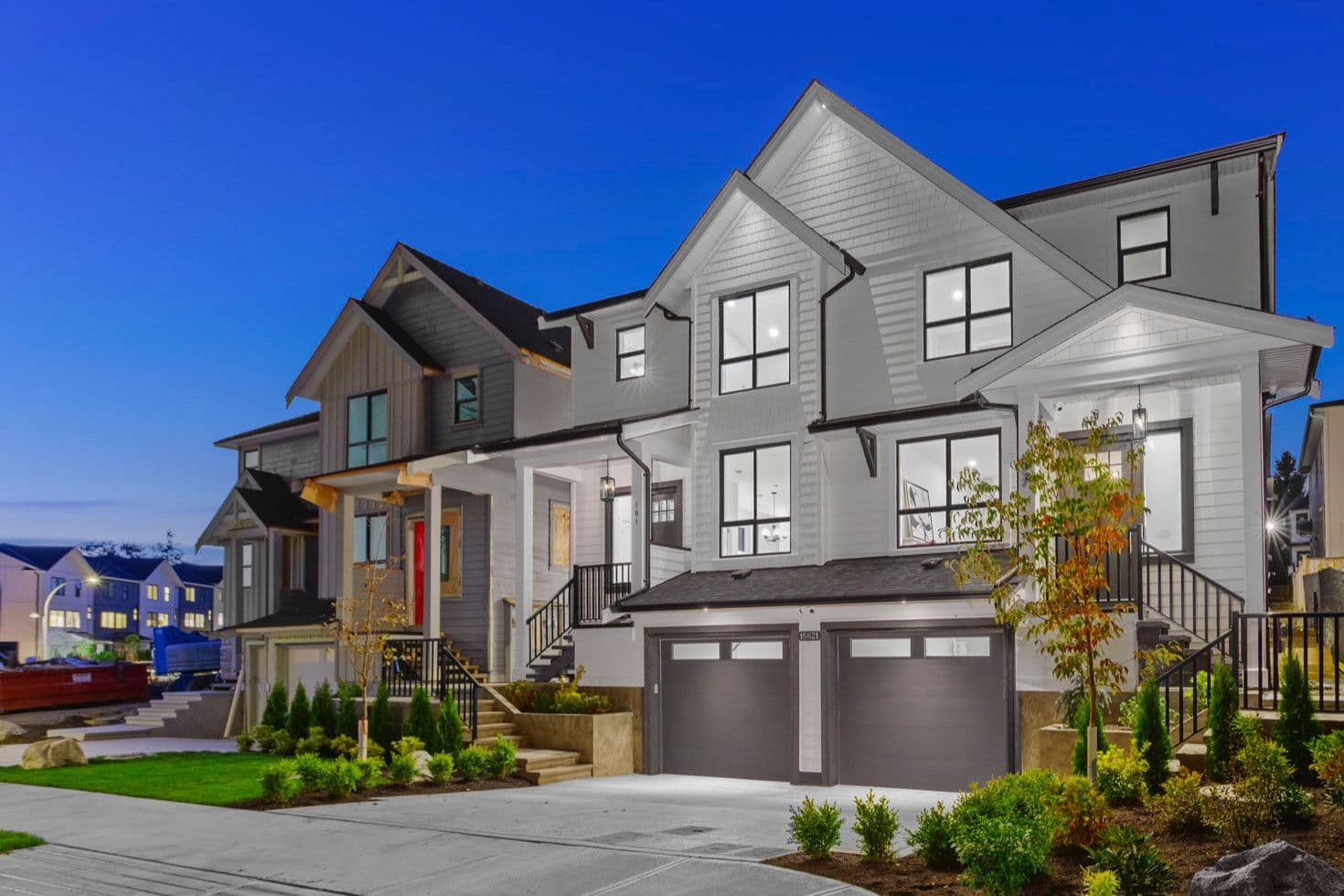 Grandview Walk – A twilight street-level view of newly constructed multi-story townhomes featuring white and grey siding, dark garage doors, and illuminated windows. Landscaped front yards with small trees, shrubs, and concrete driveways line the modern residential facades under a clear dusk sky.