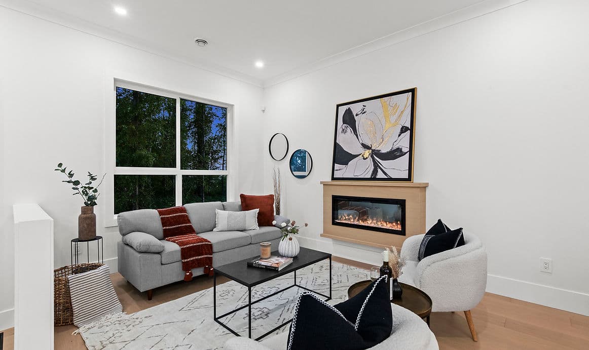 Grandview Walk – A modern living room featuring a light grey sofa, a sleek black coffee table, two textured white armchairs, and a built-in horizontal fireplace set below framed artwork. Large windows overlook lush green trees under an evening sky, complementing the clean white walls and light wood flooring.