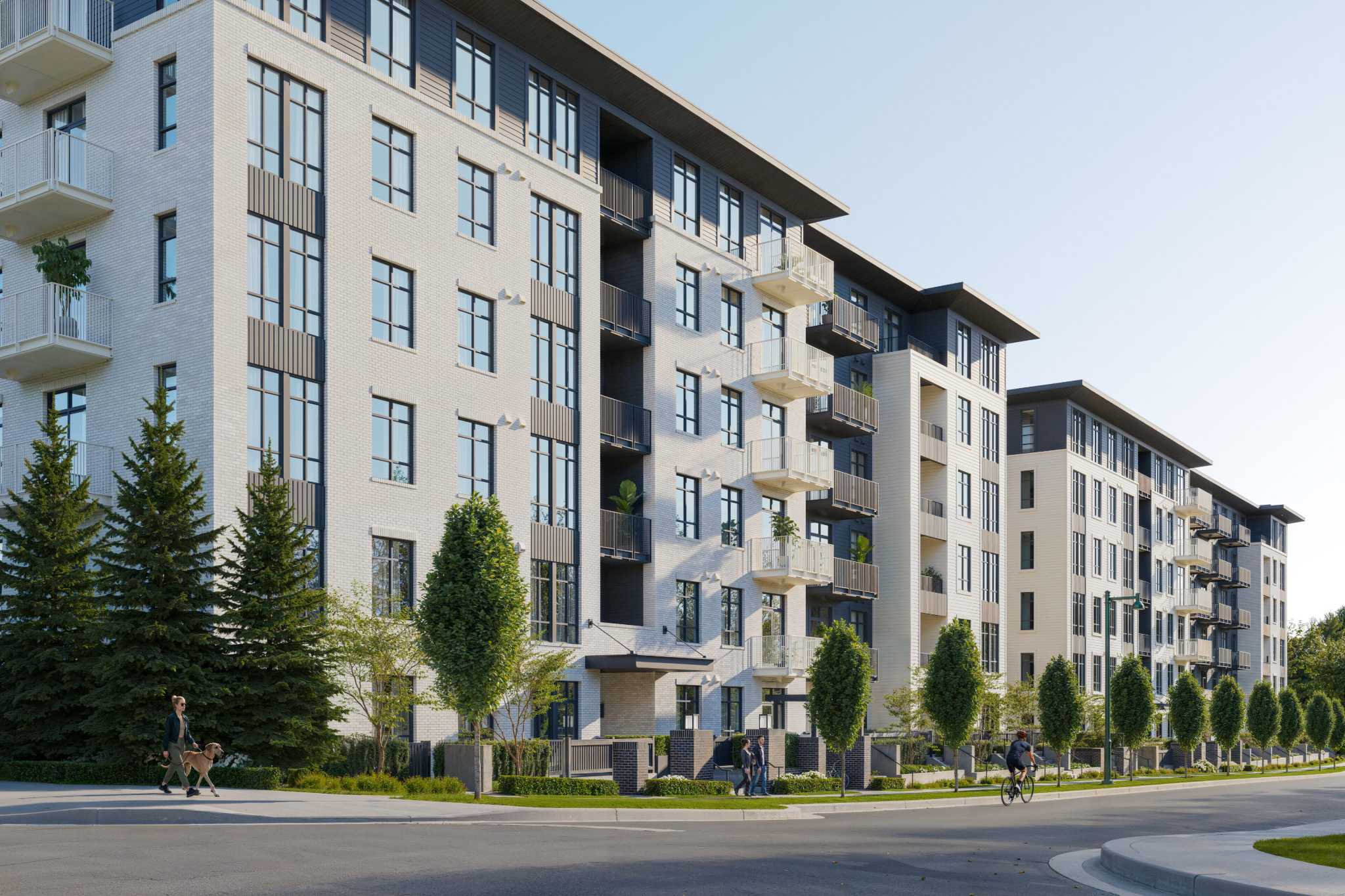 Grayson – A ground-level street view of a modern multi-story residential building featuring light grey brick, dark accent cladding, and private balconies. Pedestrians walk on a tree-lined sidewalk and a cyclist rides on the paved street beside manicured lawns under a clear blue sky.