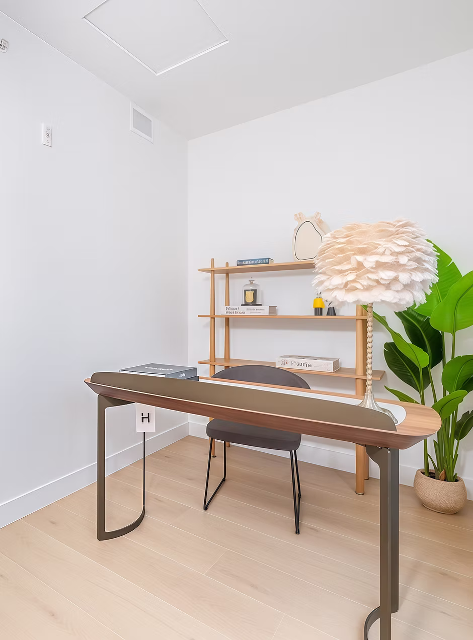 8899 Spires – A modern home office setup featuring a sleek wooden desk with a grey chair, light hardwood floors, and plain white walls. Against the back wall sits a wooden shelving unit decorated with books and trinkets, alongside a large potted green plant and a unique feathered table lamp.