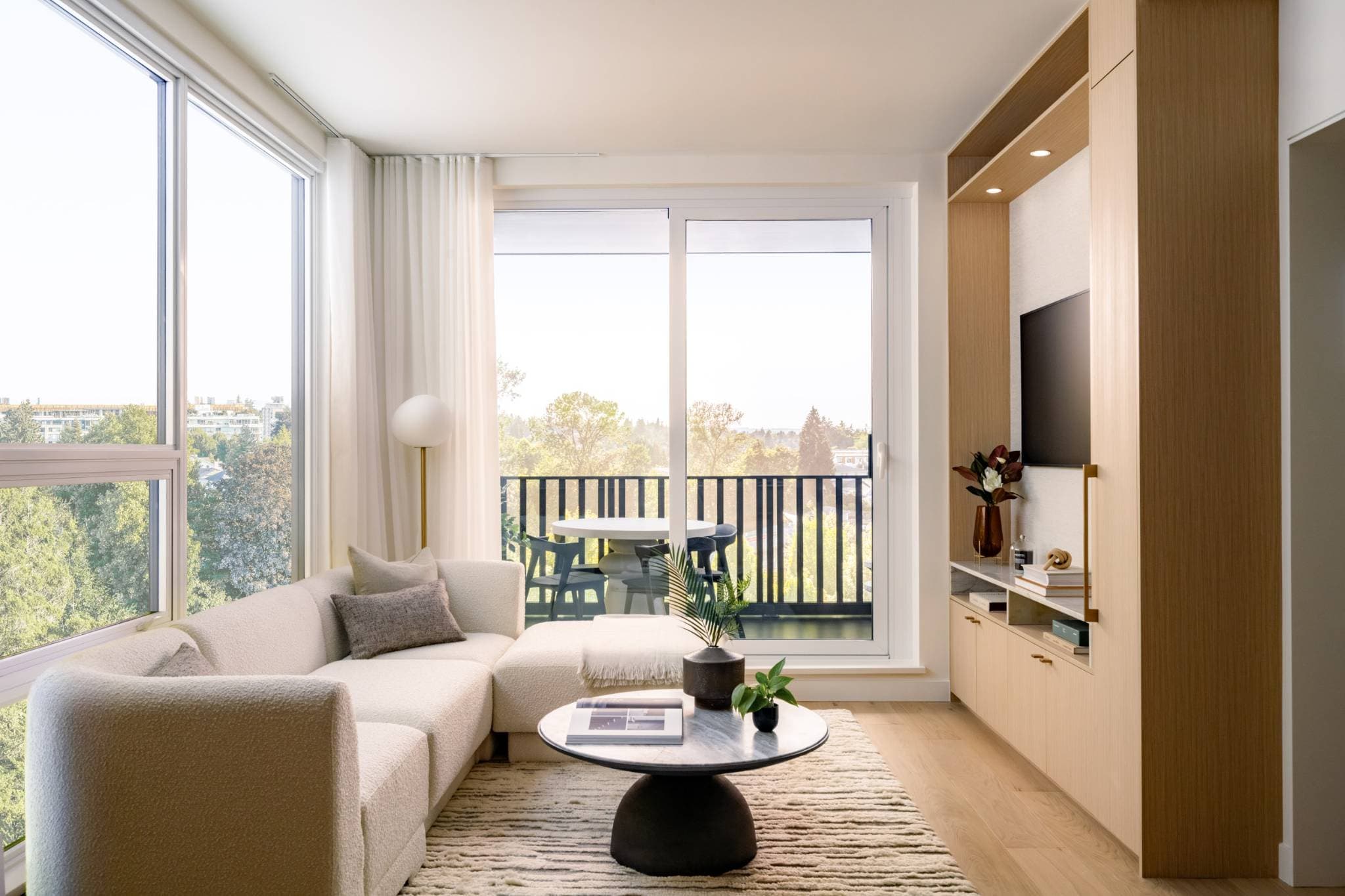 Ashleigh Oakridge Condos – Living room photo with a cream sectional sofa, wood built-in TV wall, and a balcony overlooking treetops.