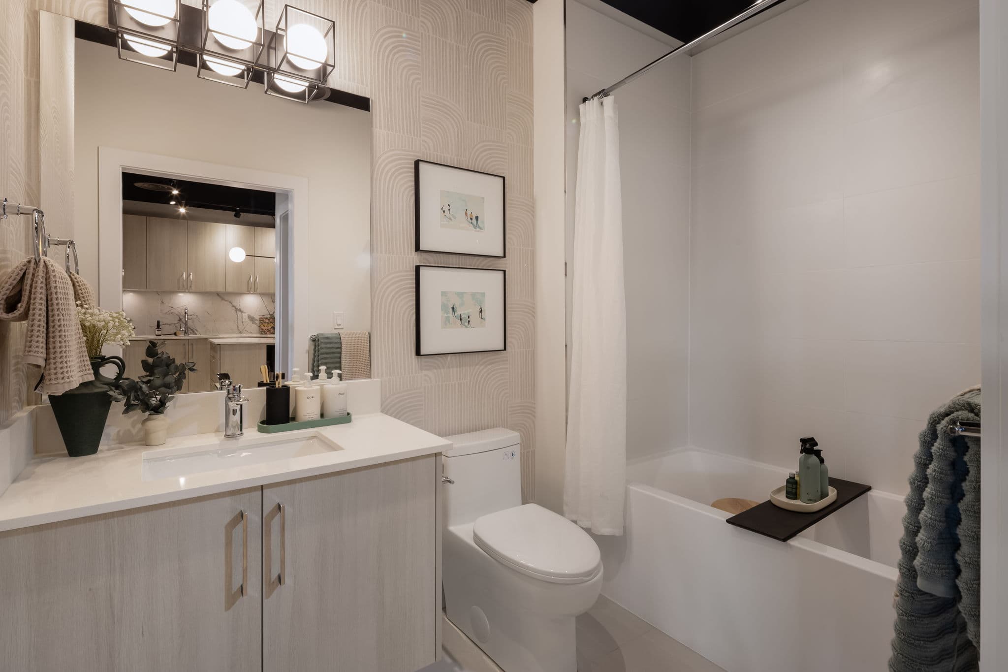 Interchange – Bathroom with a tub/shower combo behind a curtain, a vanity with framed art above the toilet, and a mirror reflecting the kitchen beyond.
