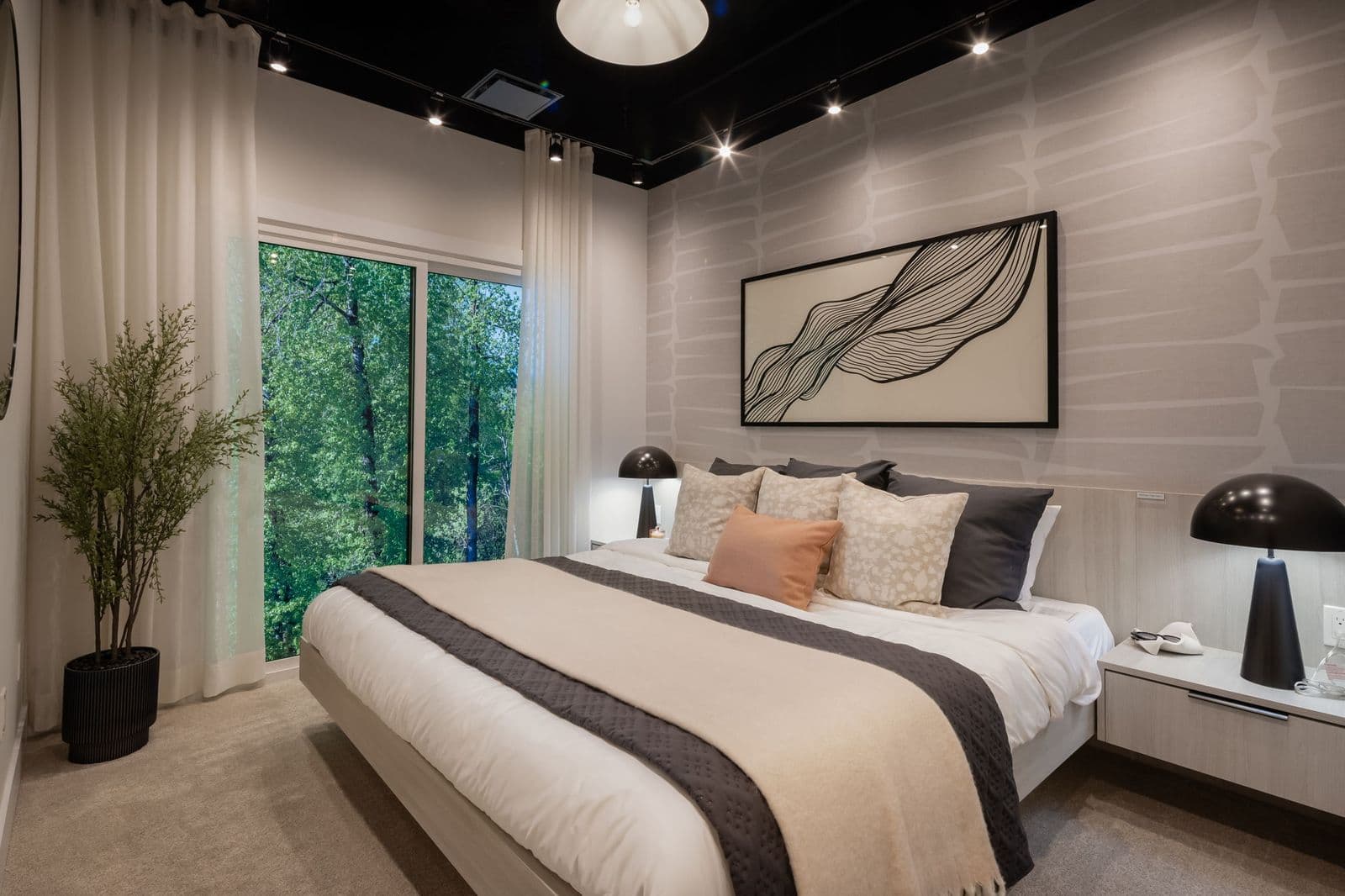 Interchange – Bedroom with a black ceiling and track lighting, patterned wallpaper, framed abstract art, a plant, and a large window looking out to trees.
