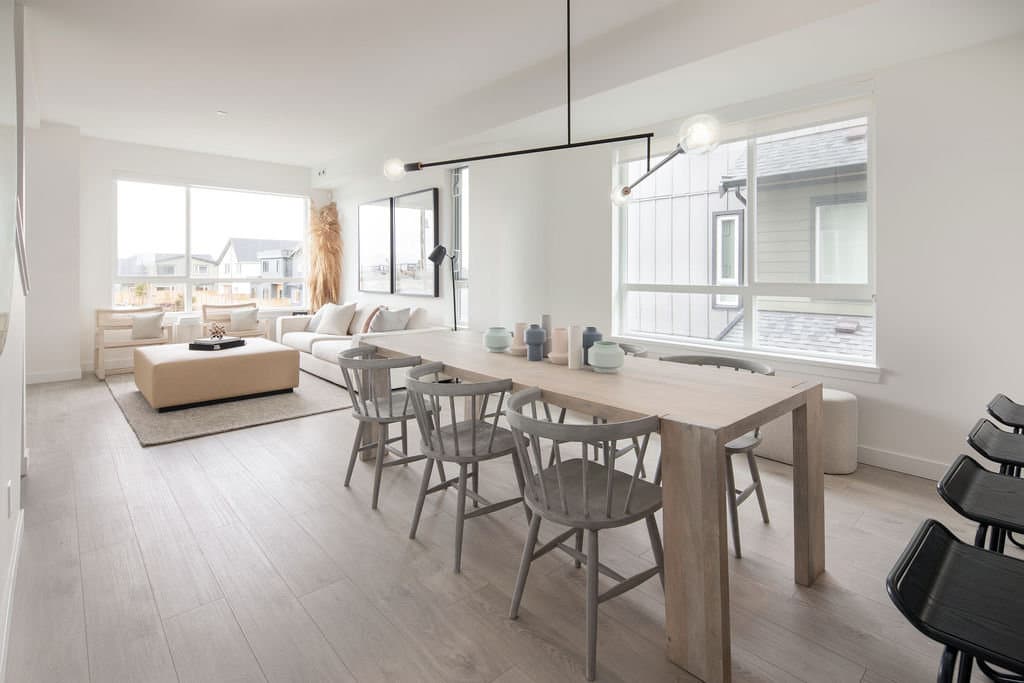 Boardwalk 6 – A bright, open-concept dining and living area featuring light wood floors, crisp white walls, and large windows that let in abundant daylight. A long wooden dining table with grey wooden spindle chairs sits under a modern black chandelier, seamless flowing into a lounge area equipped with a beige sofa, ottoman, and large window overlooking a residential neighborhood.