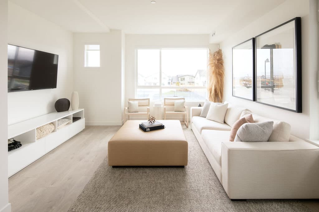 Boardwalk 6 – A bright, neutral-toned living room features a long white sofa, a large tan leather ottoman coffee table, and light wood flooring illuminated by a large back window. A mounted flat-screen TV hangs opposite framed black-and-white art prints, creating a clean, modern aesthetic.