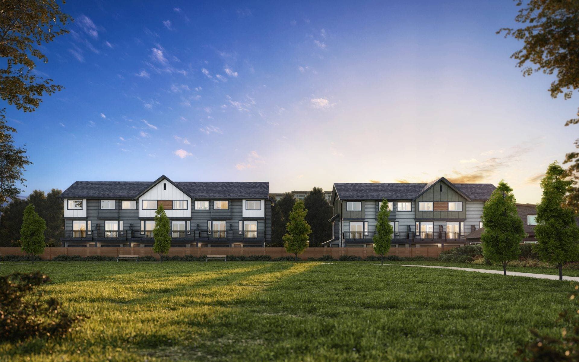 Boardwalk 6 – A ground-level exterior view shows modern three-story townhomes with dark siding and pitched roofs set behind a wooden fence. In the foreground, a expansive grassy lawn with a paved walking path and young planted trees is bathed in soft golden sunlight under a clear blue sky.