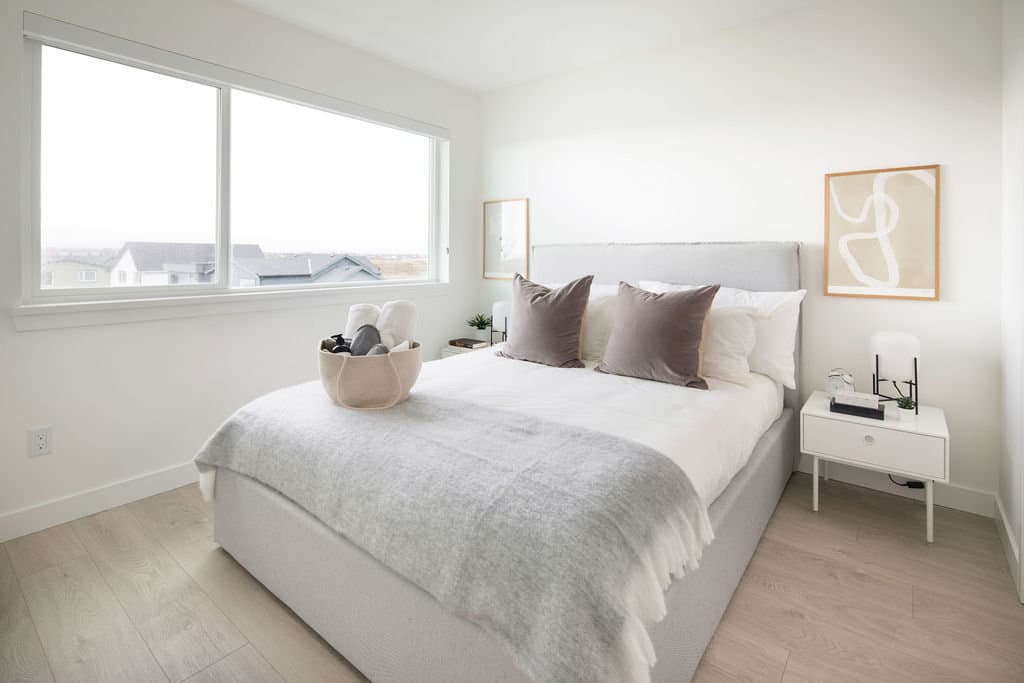 Boardwalk 6 – A bright modern bedroom staged with a grey upholstered bed, white bedding, dark grey accent pillows, and light laminate flooring. A large window overlooks neighboring rooftops under an overcast daytime sky.