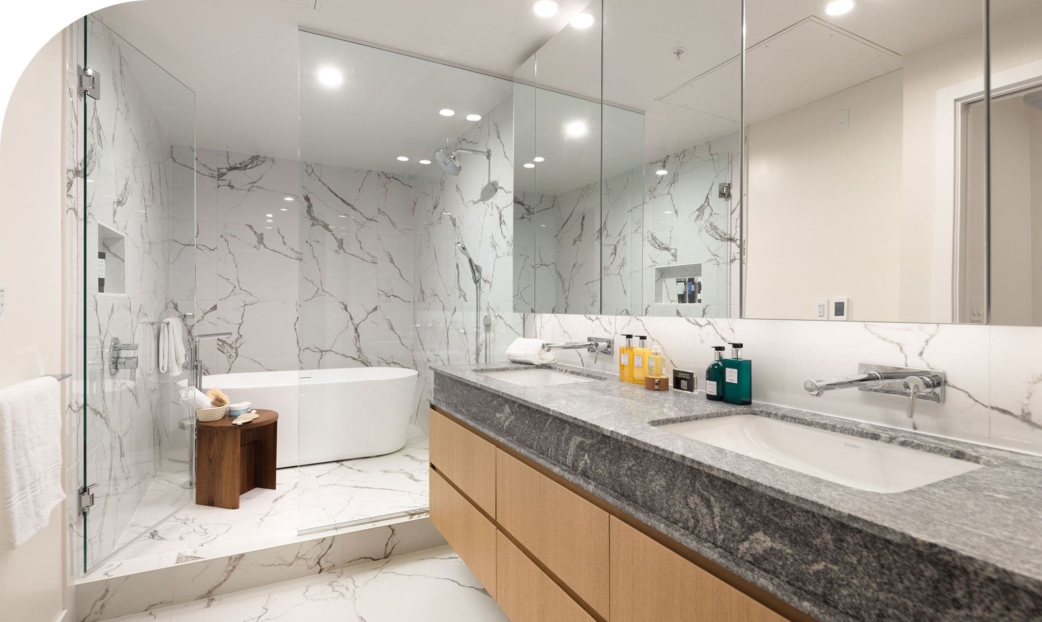 Raphael – Ensuite bathroom with a freestanding soaking tub, marble-tiled walk-in shower, and a double vanity with grey stone countertop.