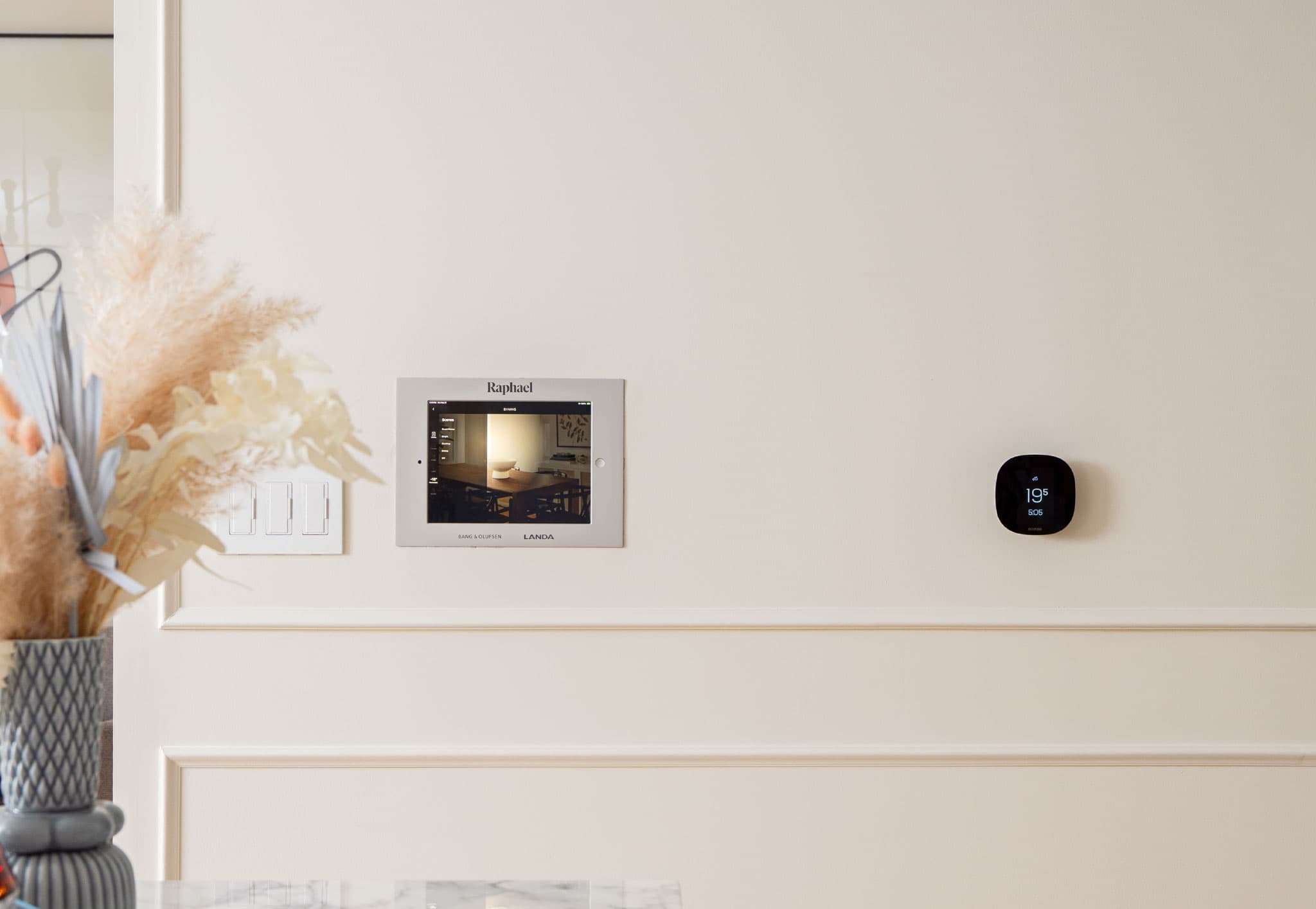 Raphael – Close-up of an in-suite smart home touchscreen control panel labeled Raphael and a wall-mounted thermostat.