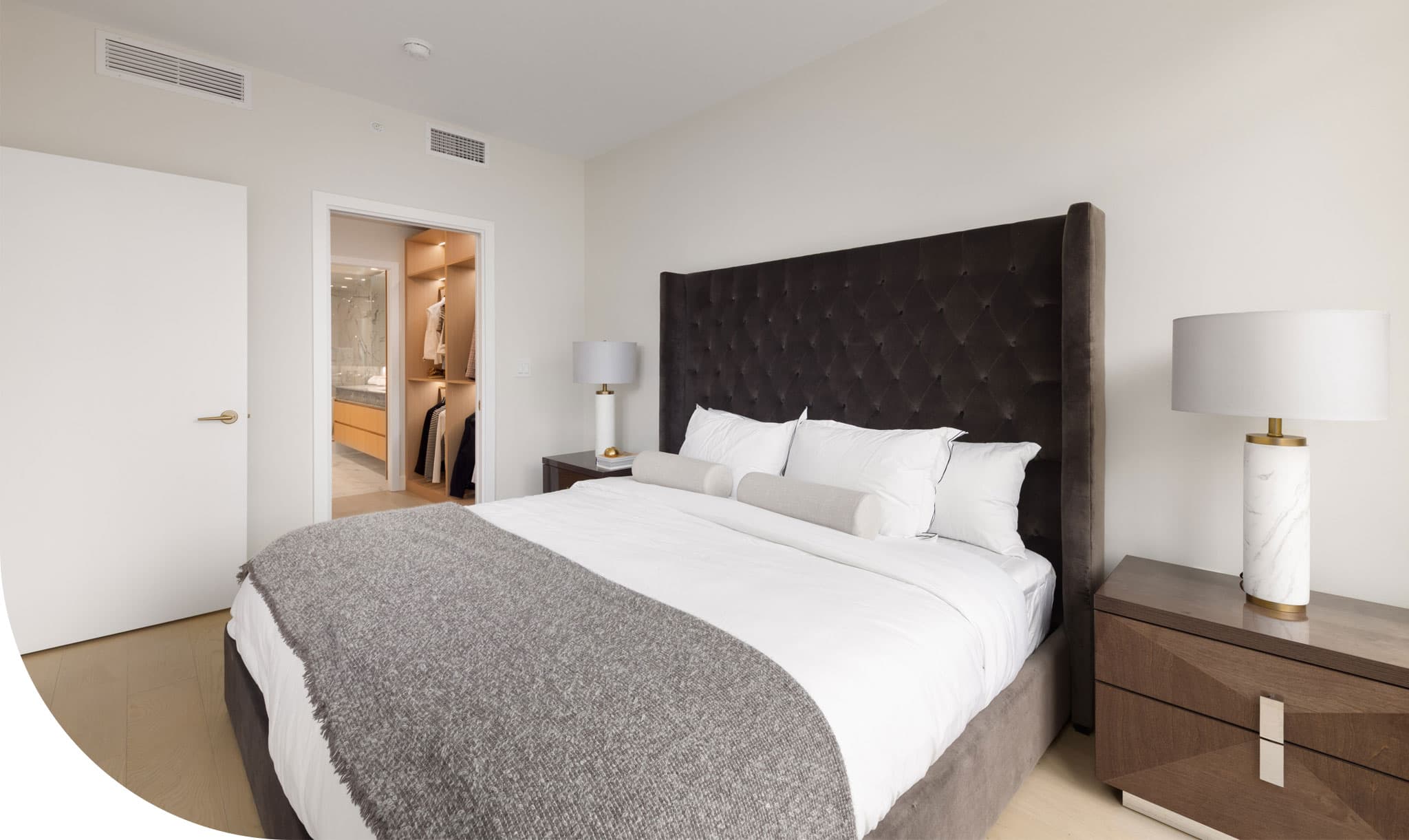 Raphael – Bedroom with a tall tufted dark headboard, white bedding, matching nightstands with marble lamps, and a view into a walk-in closet.