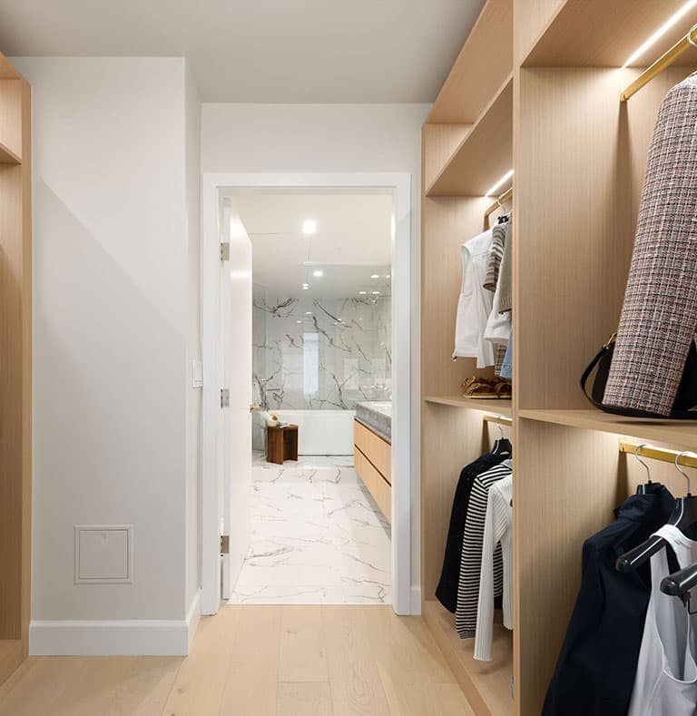 Raphael – Walk-in closet with wood shelving and hanging clothes, with an open door revealing a marble-tiled ensuite bathroom.