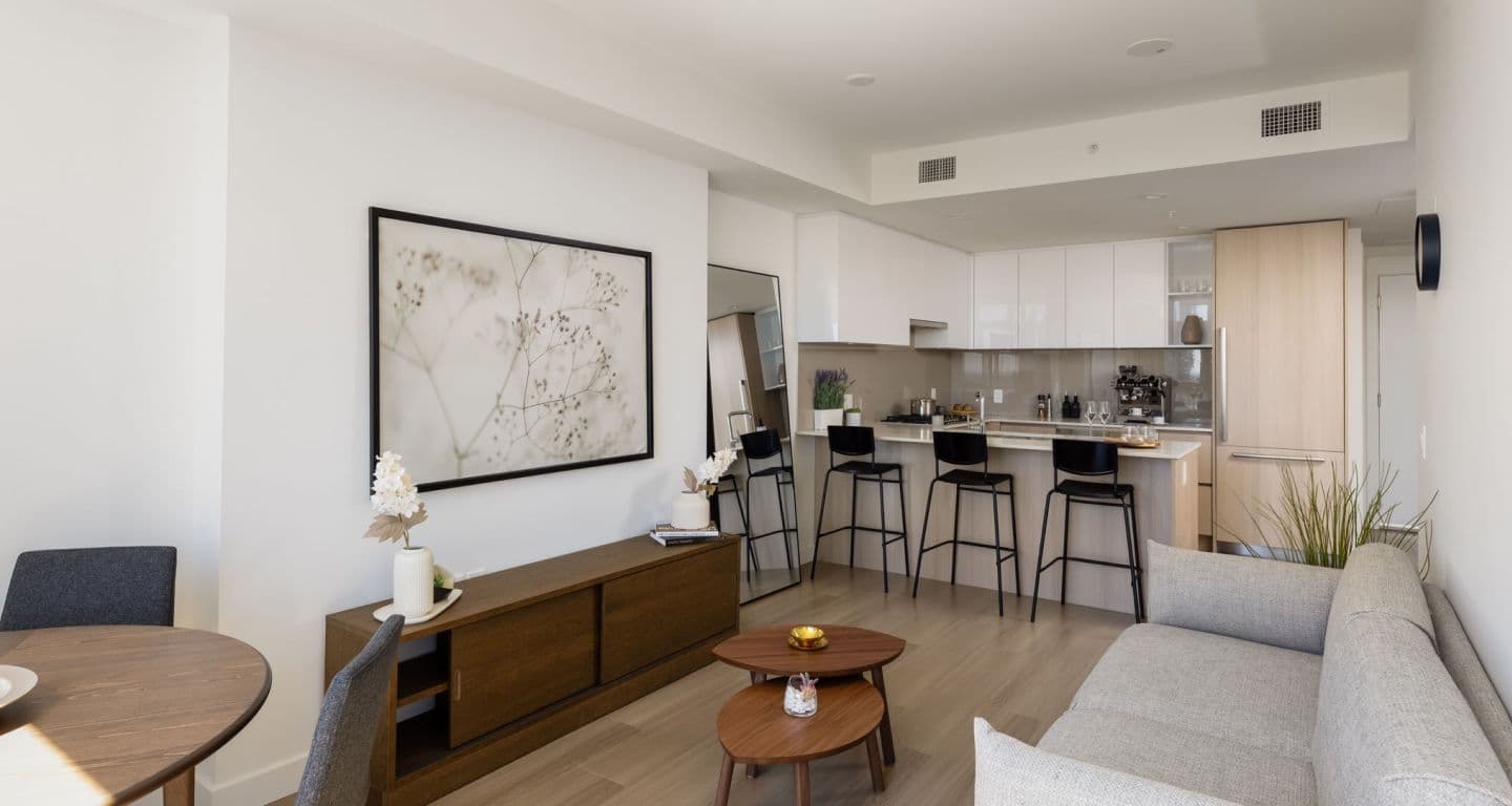 618 Carnarvon – Open living room and kitchen with a grey sofa, dark wood console table, framed botanical art, and a white kitchen with a breakfast bar and four black stools.