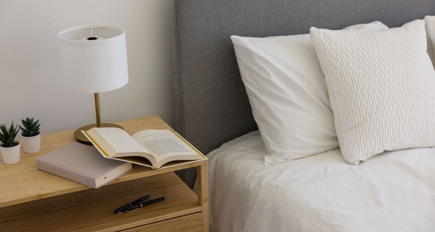 618 Carnarvon – Close-up of a bedroom nightstand with a lamp, small succulents, an open book, and a corner of a white-bedded bed with a grey upholstered headboard.