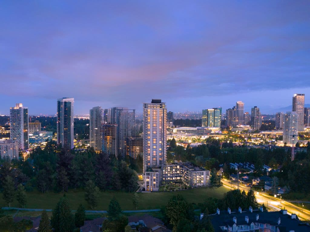 SkyLiving – A dusk aerial photo of the same lit-up high-rise tower among other towers, trees, and a residential neighbourhood with a busy roadway below.
