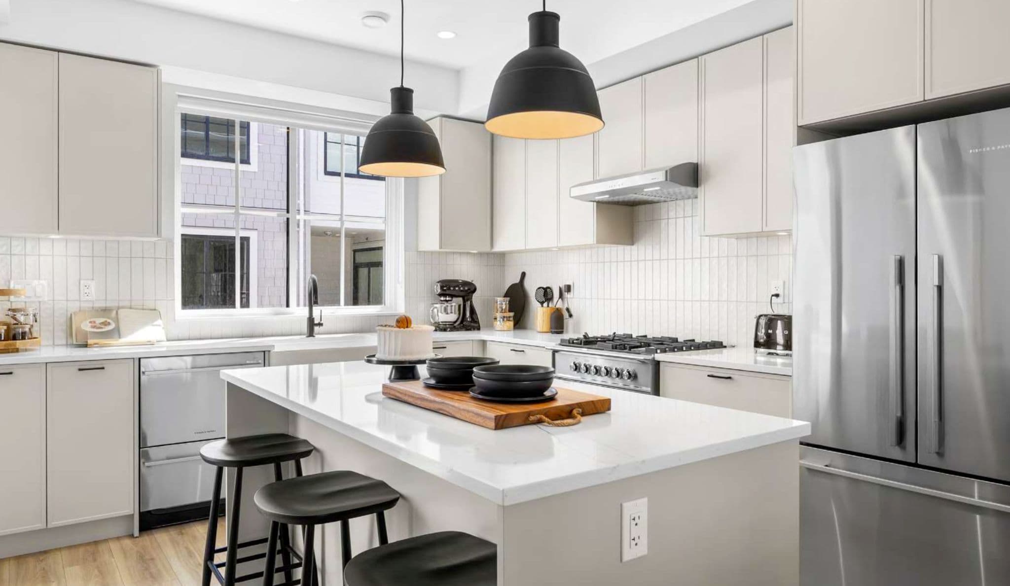 Heath West – Kitchen with light cabinetry, a large island with black stools, black pendant lights, and a stainless steel refrigerator.