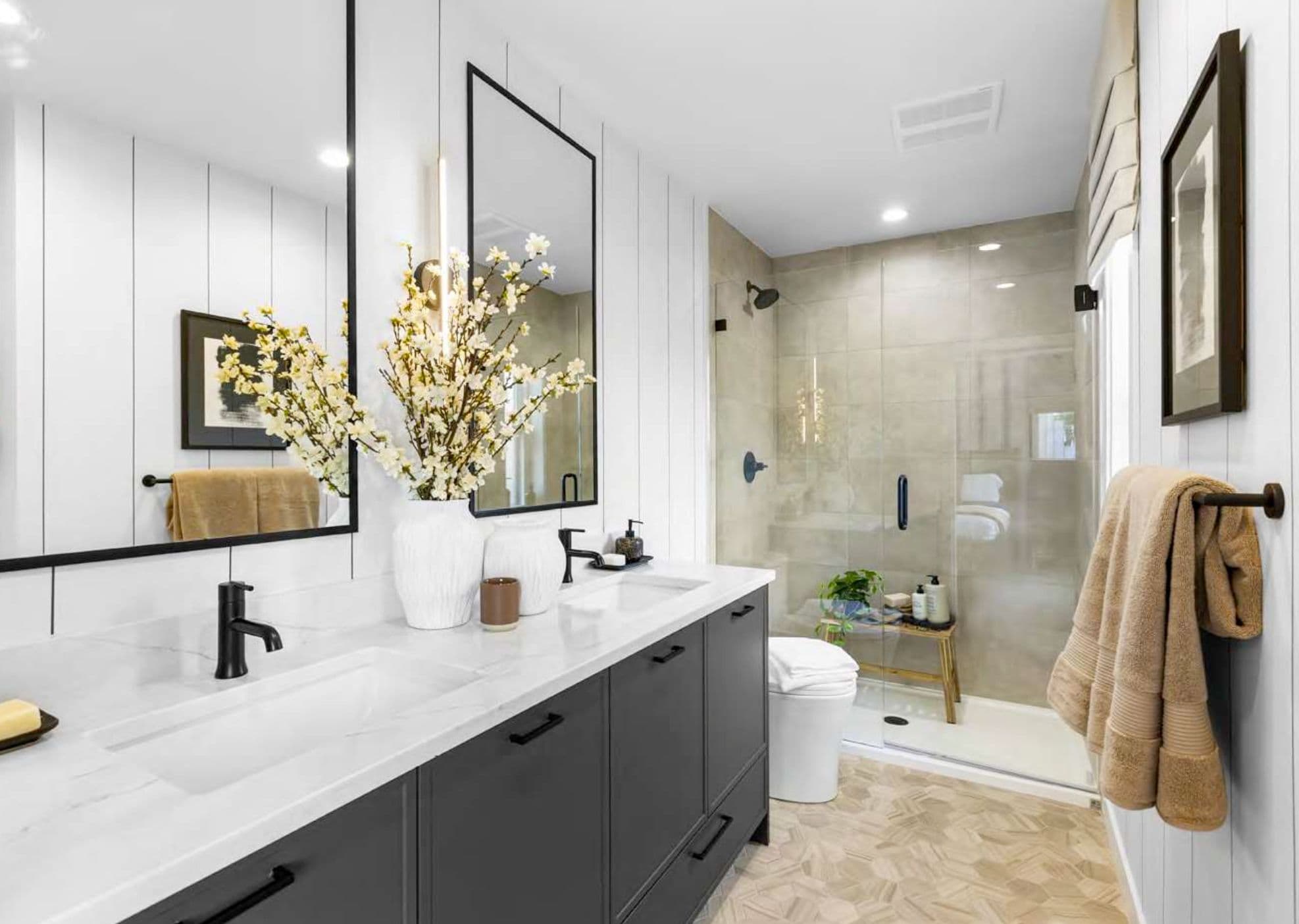 Heath West – Bathroom with dark vanity cabinets, white marble-look countertop, and a glass shower stall with tiled walls.