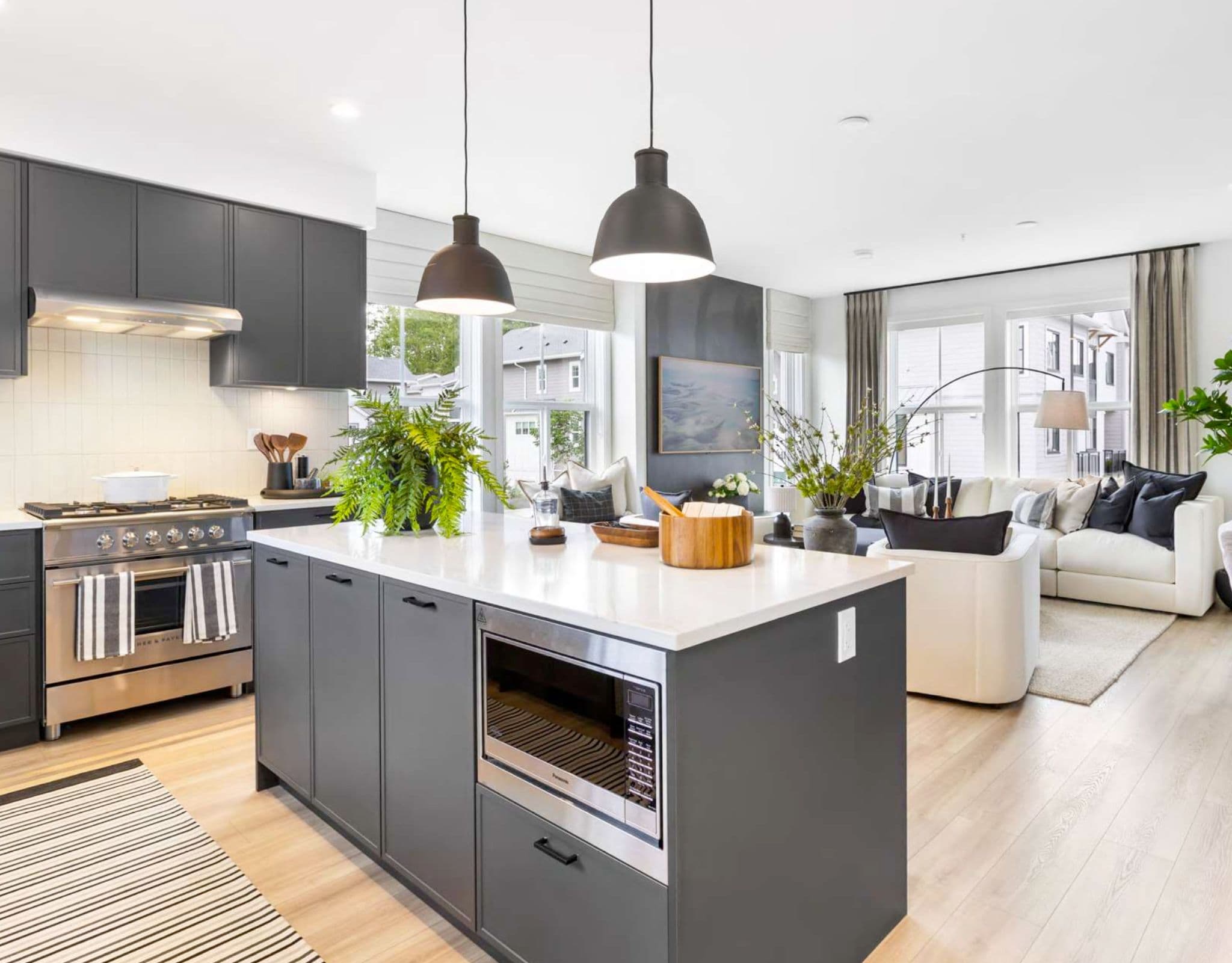 Heath West – Open-concept kitchen with dark cabinetry and island opening onto a bright living room with a white sofa.