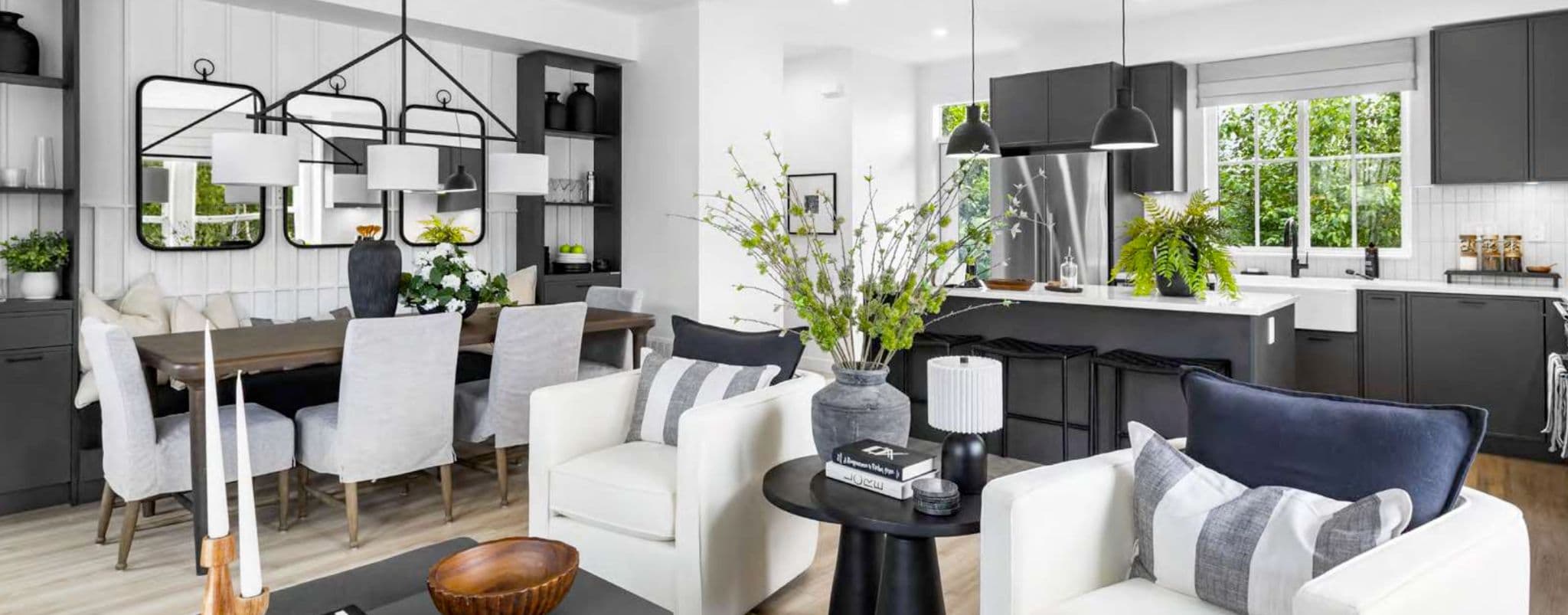 Heath West – Wide view of an open-concept dining table, living room seating area, and kitchen island with dark cabinetry in the background.