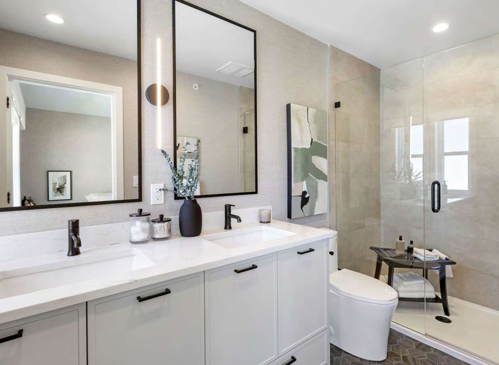 Heath West – Bathroom with a long double-sink vanity, black-framed mirrors, and a glass-enclosed shower with a small bench seat.