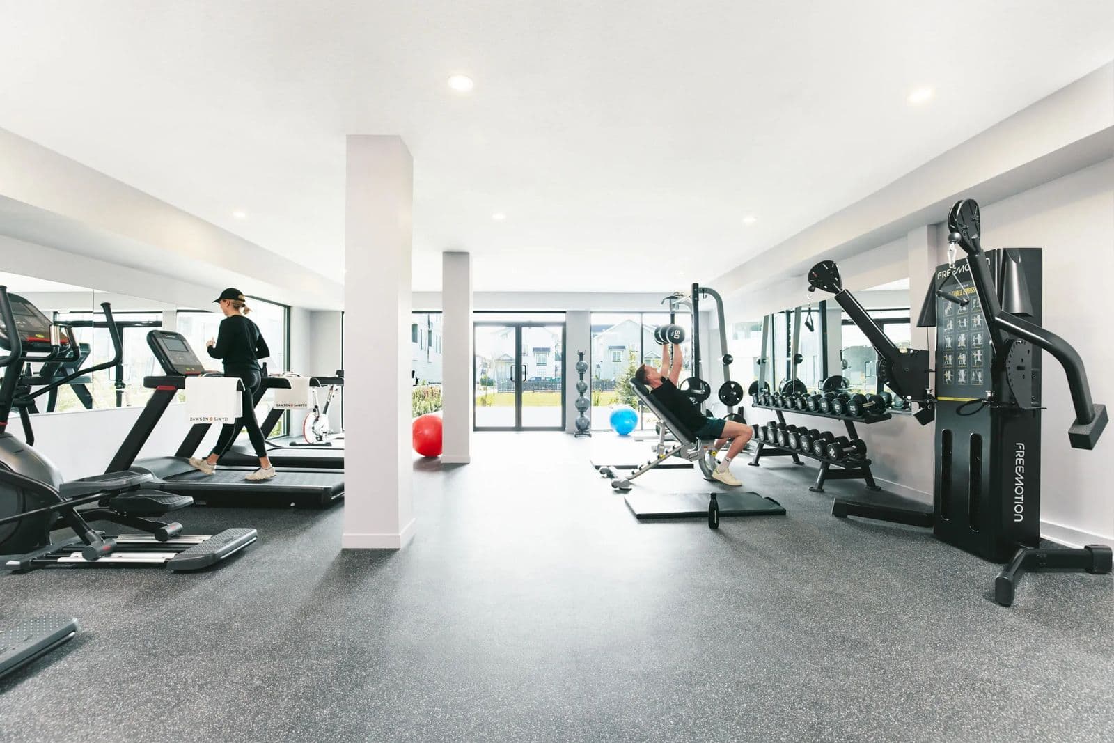 Fleetwood Village 2 – Bright fitness room with treadmills, ellipticals, a weight bench, dumbbell rack, and cable machine; a person runs on a treadmill and another lifts weights near floor-to-ceiling windows.