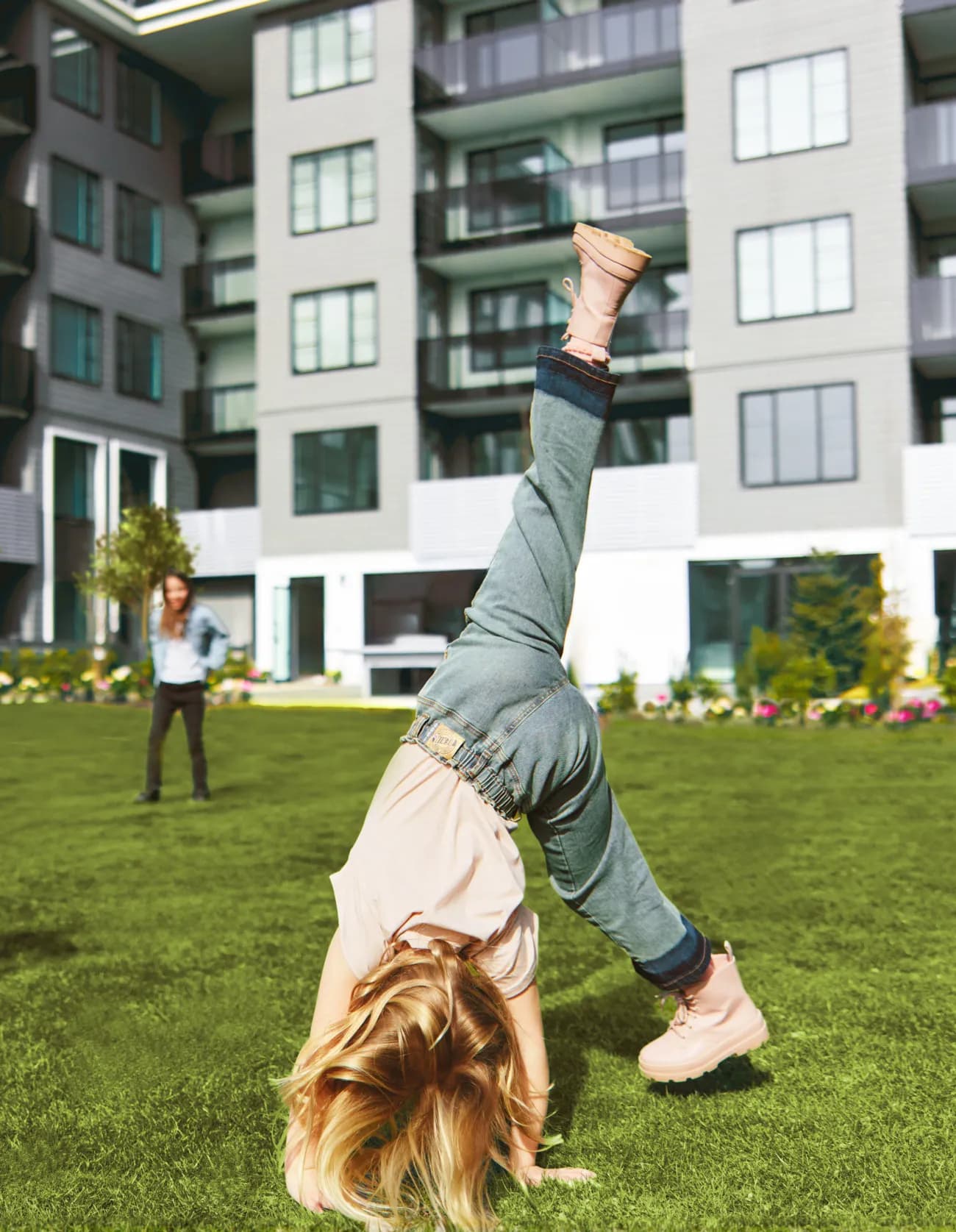 Fleetwood Village 2 – A child does a cartwheel on a landscaped courtyard lawn in front of the mid-rise residential building, with flower beds and another person watching.
