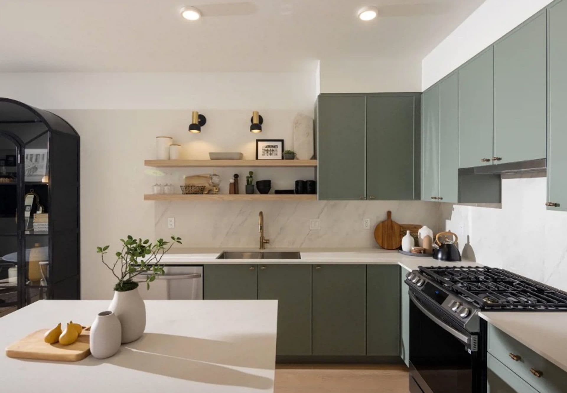 Bastille – Kitchen with sage green cabinetry, brass hardware, open wood shelving, a marble backsplash, and a gas range beside a black arched display cabinet.