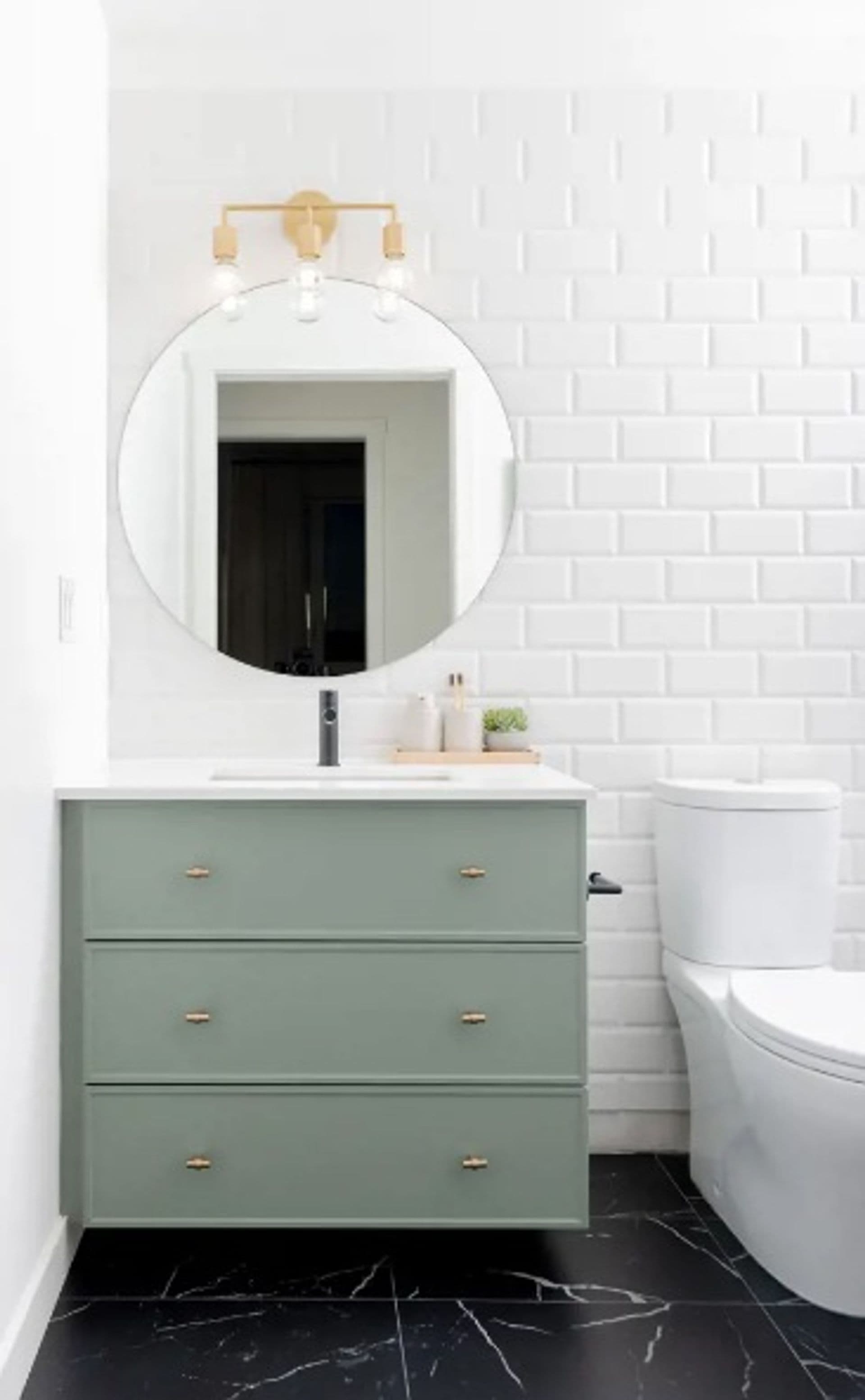 Bastille – Bathroom with white subway tile, a round mirror with brass sconce, sage green three-drawer vanity, and a white toilet on black marble-look flooring.