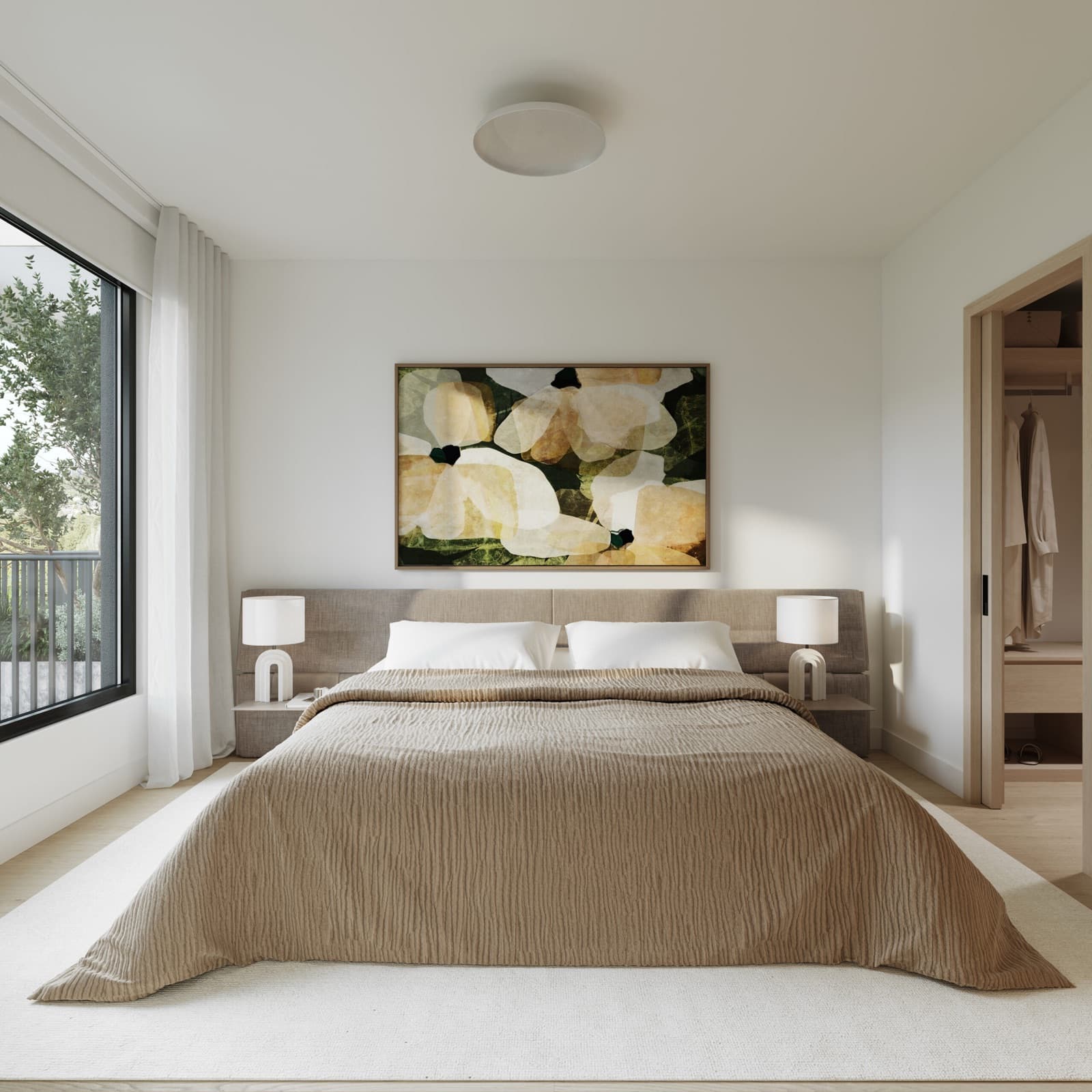 Siena – A bedroom with a large bed dressed in a taupe textured coverlet, a padded upholstered headboard, matching nightstand lamps, a floral art print, and a walk-in closet visible through a doorway.