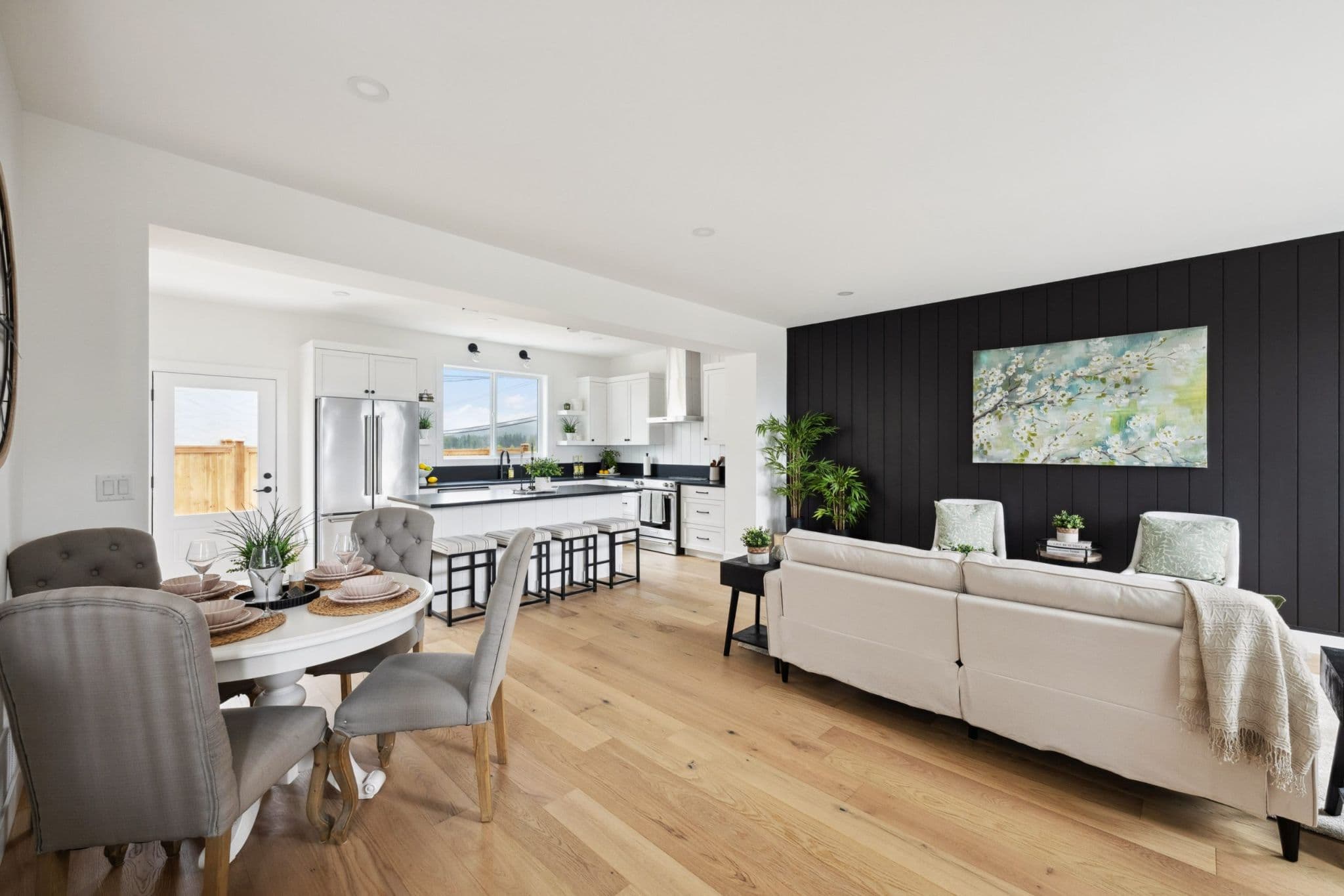 Water Tower Hill – A bright, open-concept living area featuring light wood flooring, a dining set with upholstered gray chairs, and a living room centered around a dark vertical shiplap accent wall. The space extends into a modern white kitchen equipped with an island, bar seating, and stainless steel appliances.
