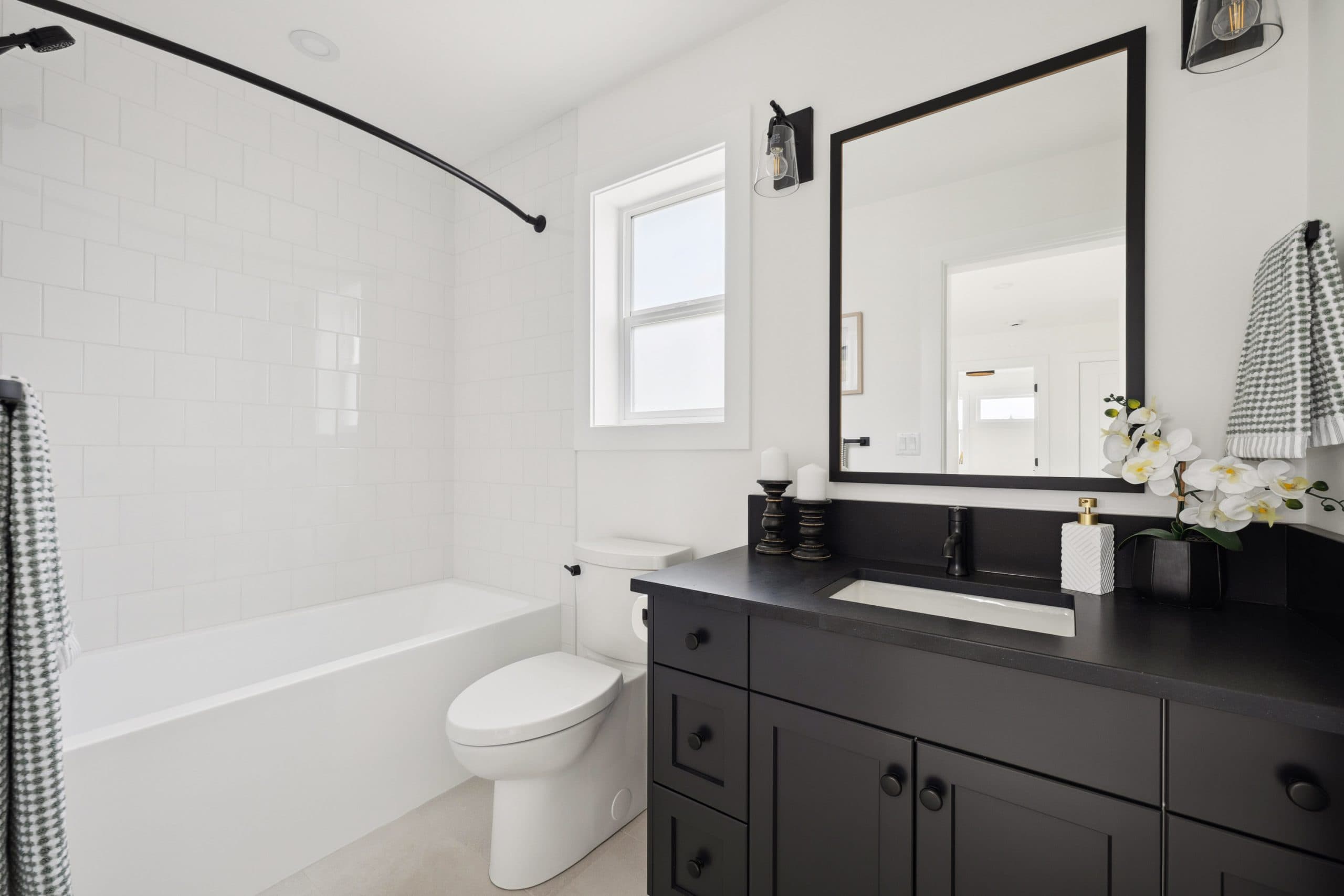 Water Tower Hill – A bright, modern bathroom featuring a black vanity with a matching countertop, sink, and matte black fixtures contrasted against white walls and white subway tile. A combination shower and bathtub lines one side beneath a frosted window, while a large black-framed mirror sits above the vanity adorned with white orchid decor.