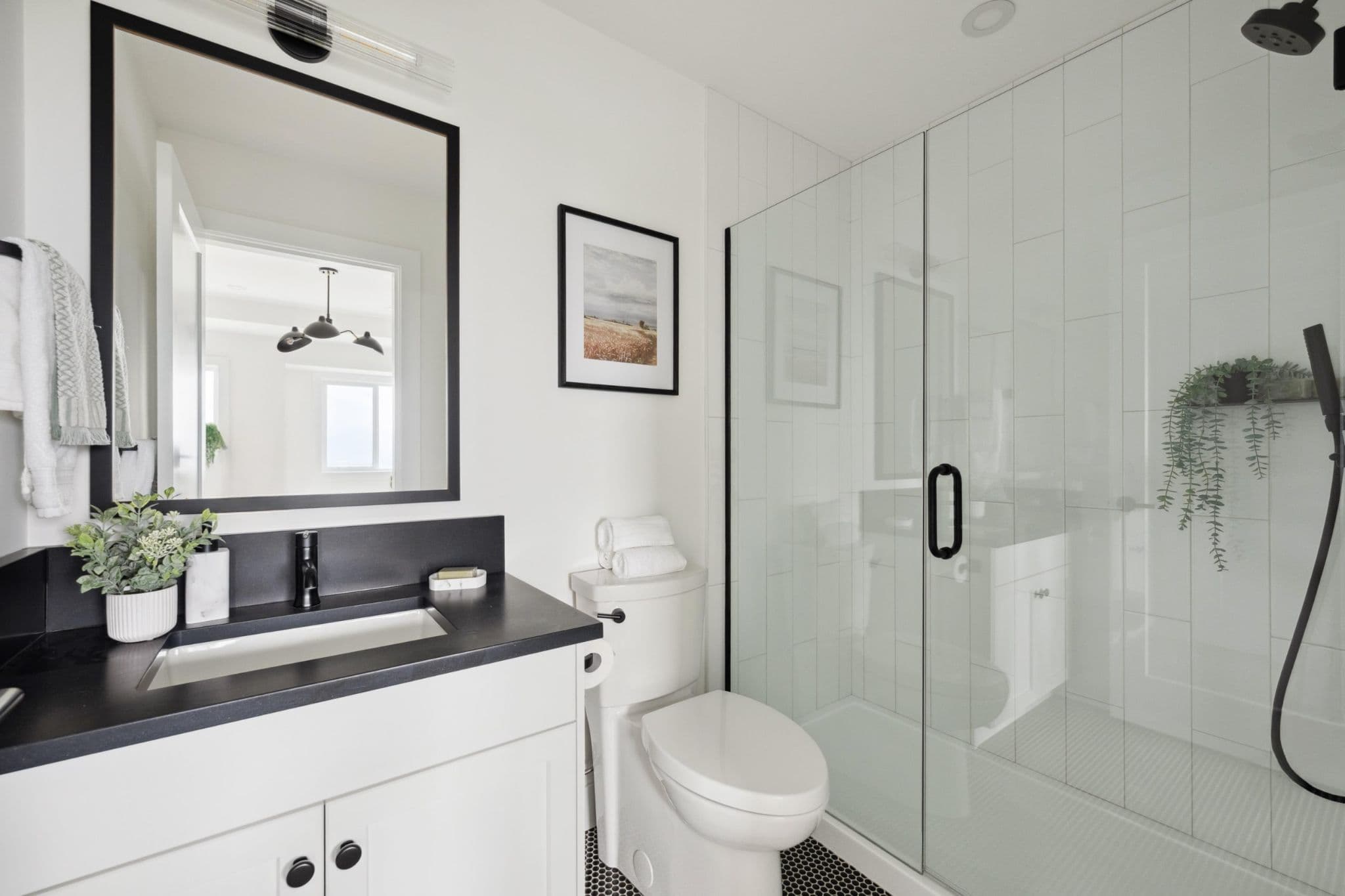 Water Tower Hill – A modern, brightly lit bathroom featuring a white vanity with a black countertop and matte black hardware, positioned beside a toilet and a glass-enclosed walk-in shower with vertical white tile. Black framed mirrors and framed artwork complement the black-and-white color palette accented with subtle potted greenery.