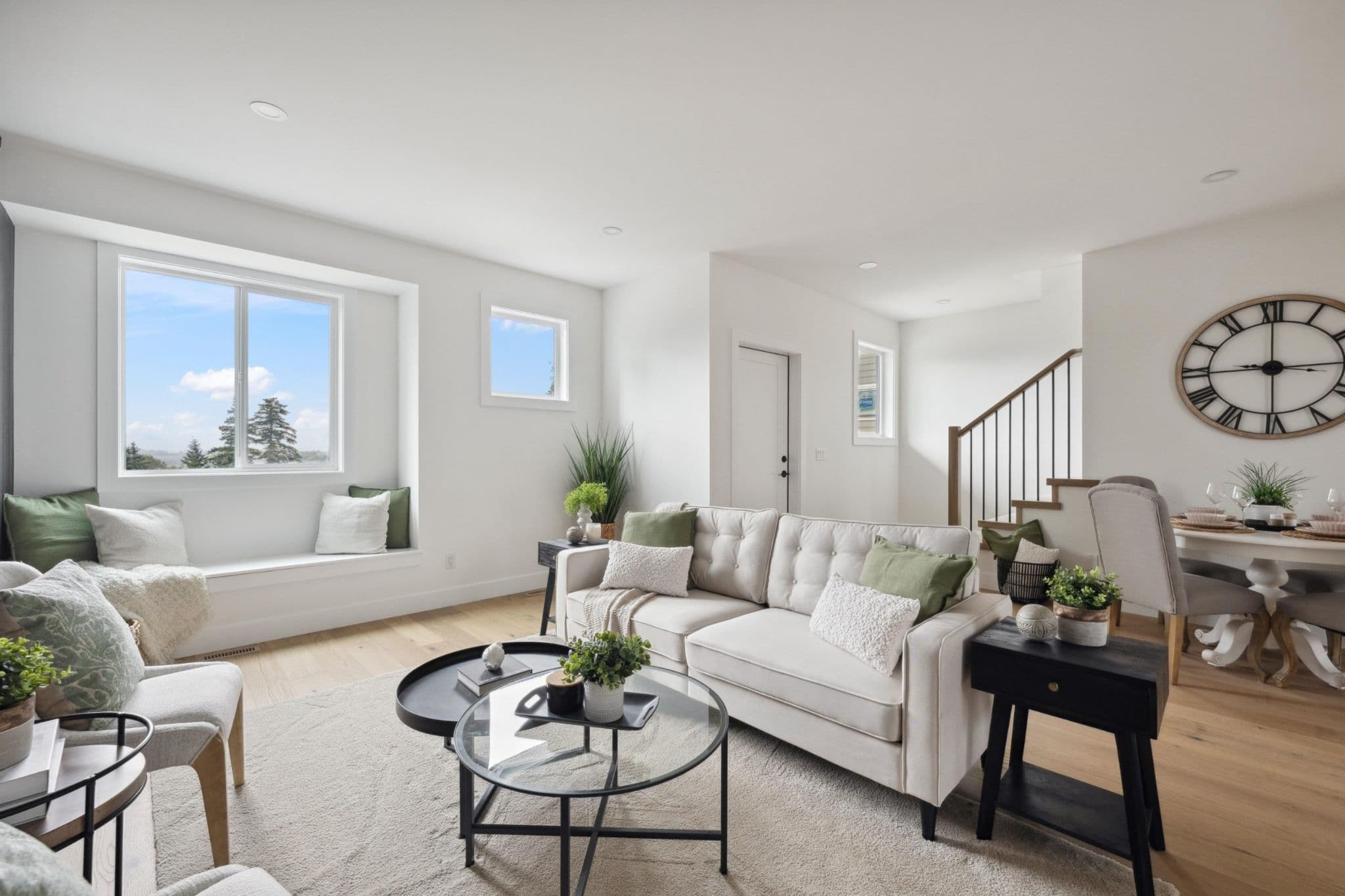 Water Tower Hill – A bright, open-concept modern living room with light hardwood floors, white walls, and a built-in window bench looking out toward distant trees and a blue sky. The space is styled with a cream tufted sofa, green accent pillows, round glass-and-metal coffee tables, and a staircase leading to an upper floor in the background.