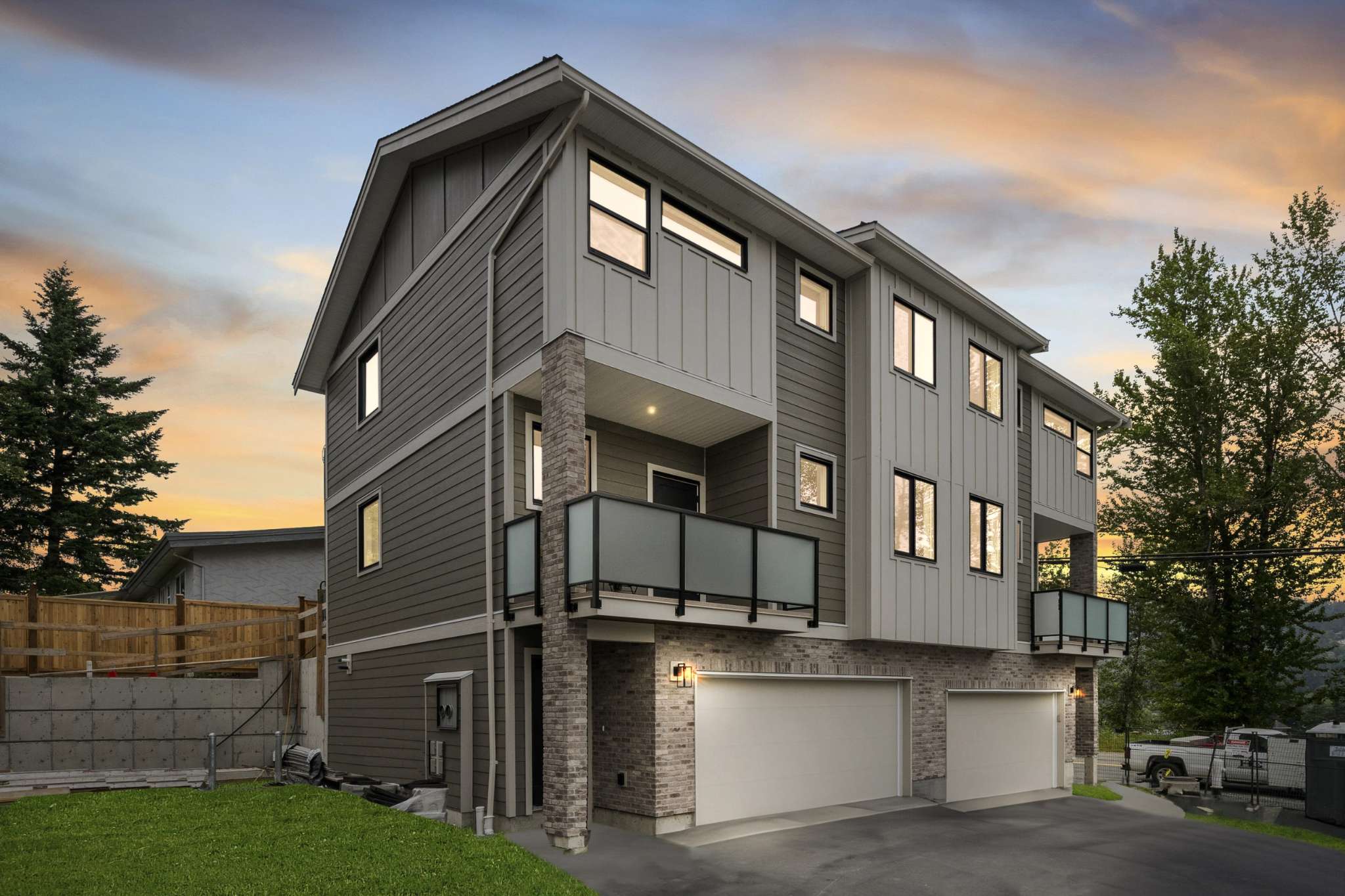 Water Tower Hill – A three-story modern townhouse features grey horizontal siding, vertical board-and-batten accents, and a brick veneer ground level with multi-car garage doors. Balconies with frosted glass railings overlook the driveway against a dramatic sunset sky framed by tall evergreen trees.