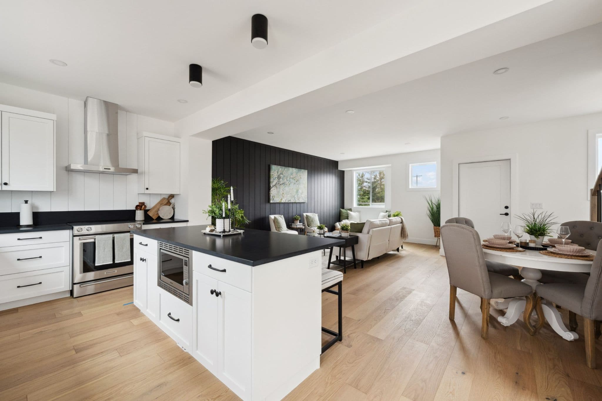 Water Tower Hill – A bright, open-concept main floor features a kitchen island with a black countertop and integrated microwave, leading into a dining area and a living room accented by a dark vertical shiplap wall. Light wood flooring runs throughout the space, complemented by crisp white cabinetry, stainless steel appliances, and abundant natural light from the living area windows.