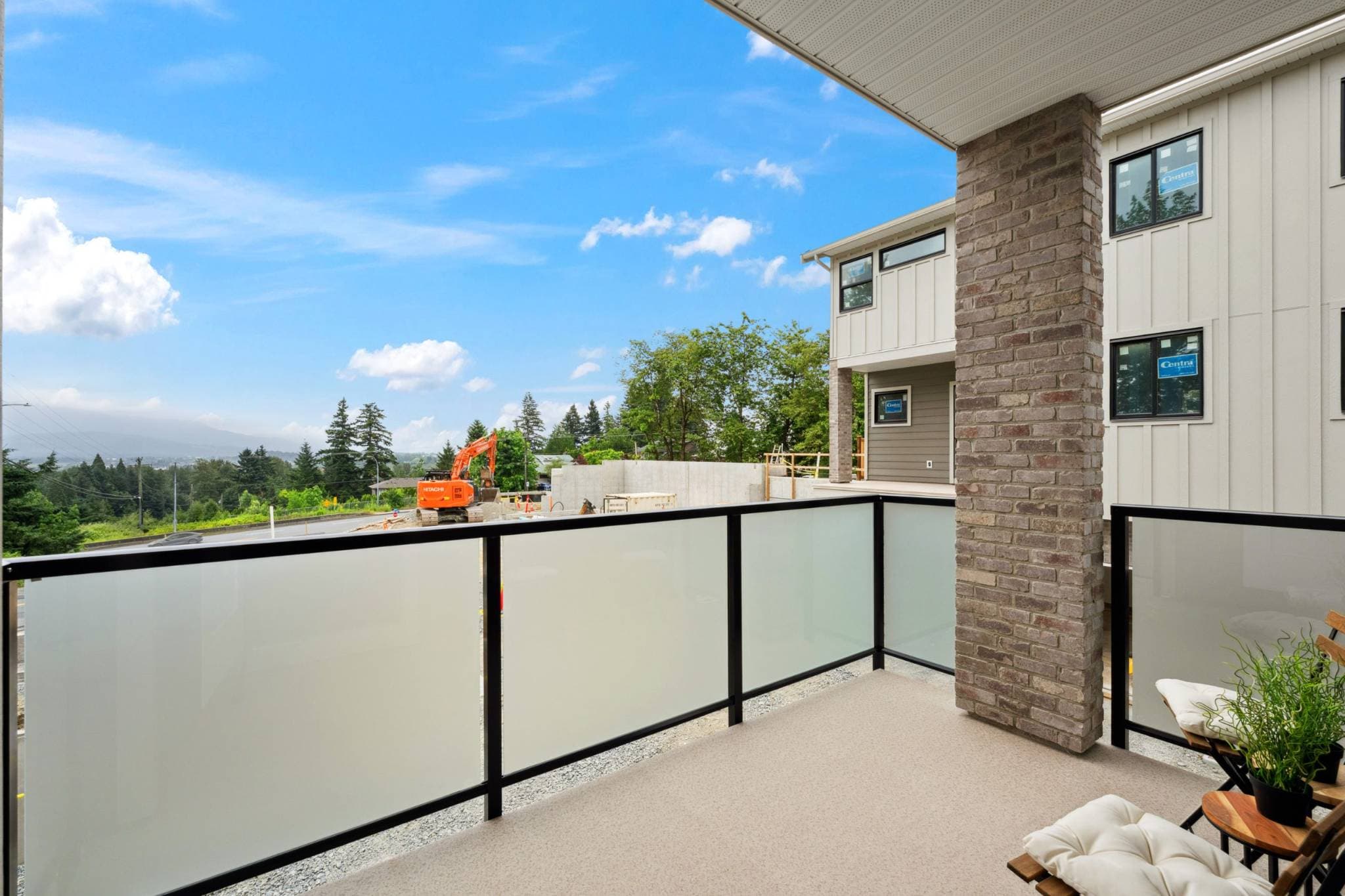 Water Tower Hill – A covered outdoor balcony features frosted-glass privacy railings, a brick-faced support pillar, and patio chairs. Beyond the railing, the daylight view overlooks an active construction site with an excavator, leading out toward lush green treetops and distant hazy mountains under a blue sky.