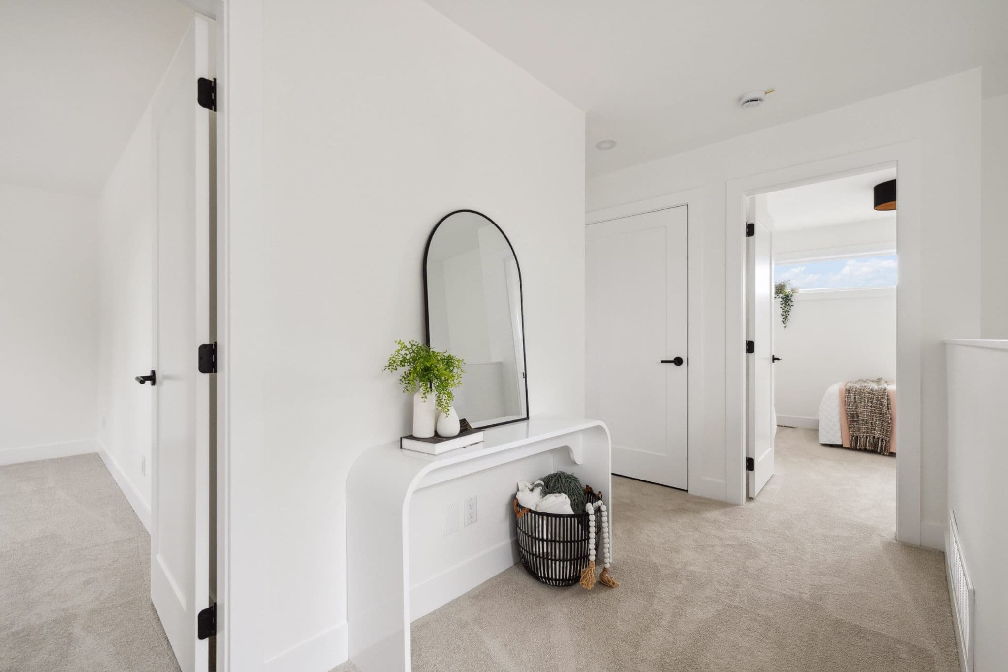 Water Tower Hill – A bright, modern interior hallway with plush light-toned carpeting and crisp white walls features an arched black-framed mirror atop a sleek curved console table. Minimalist white doors with contrasting black hardware line the space, offering a view into an adjacent sunlit bedroom.