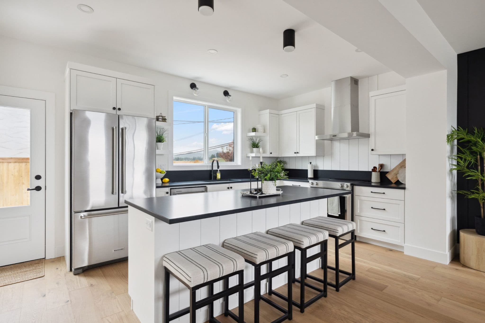Water Tower Hill – A bright, modern farmhouse-style kitchen features white shaker cabinetry, contrasting black countertops, and light wood flooring. A central island with vertical paneling and four cushioned barstools anchors the space, framed by stainless steel appliances and a window looking out toward distant hills.