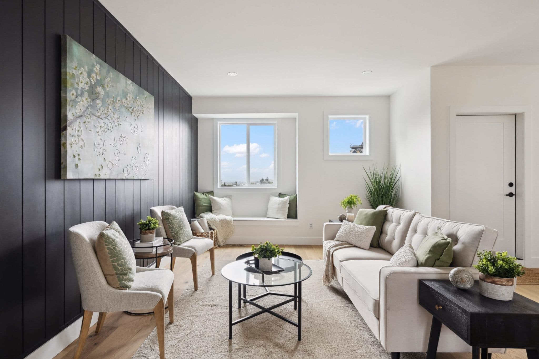 Water Tower Hill – A brightly lit, contemporary living room features a dark charcoal vertical shiplap accent wall decorated with floral artwork, contrasted by crisp white walls and light hardwood floors. The space is styled with a plush off-white sofa, two light armchairs, a glass coffee table, and a cozy built-in window bench beneath a bright window looking out toward a blue sky.