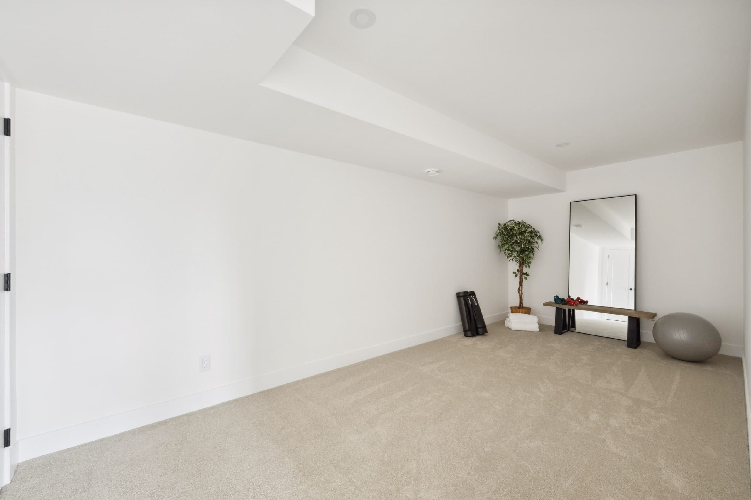Water Tower Hill – A spacious, minimalist room with beige carpeting and plain white walls, featuring a bulkhead along the ceiling. In the far corner, a small workout setup includes rolled-up yoga mats, a potted plant, a wooden bench with dumbbells, a large leaning floor mirror, and an exercise ball.
