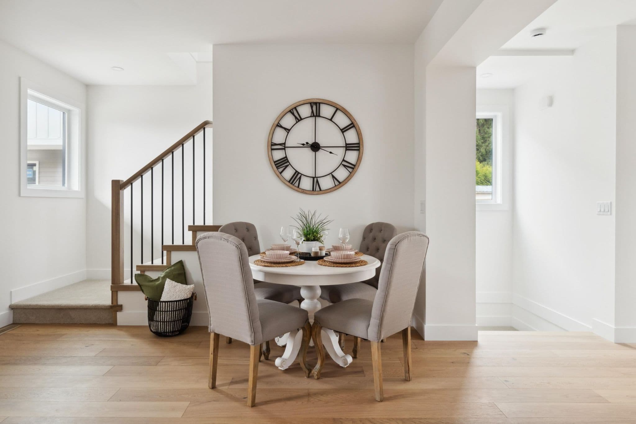 Water Tower Hill – A bright, cozy dining nook features a round white pedestal table set for four with upholstered beige chairs, light wood flooring, and a large decorative wall clock above. Adjacent to the dining space is a staircase with black metal balusters and natural wood railings leading to the upper level of the home.