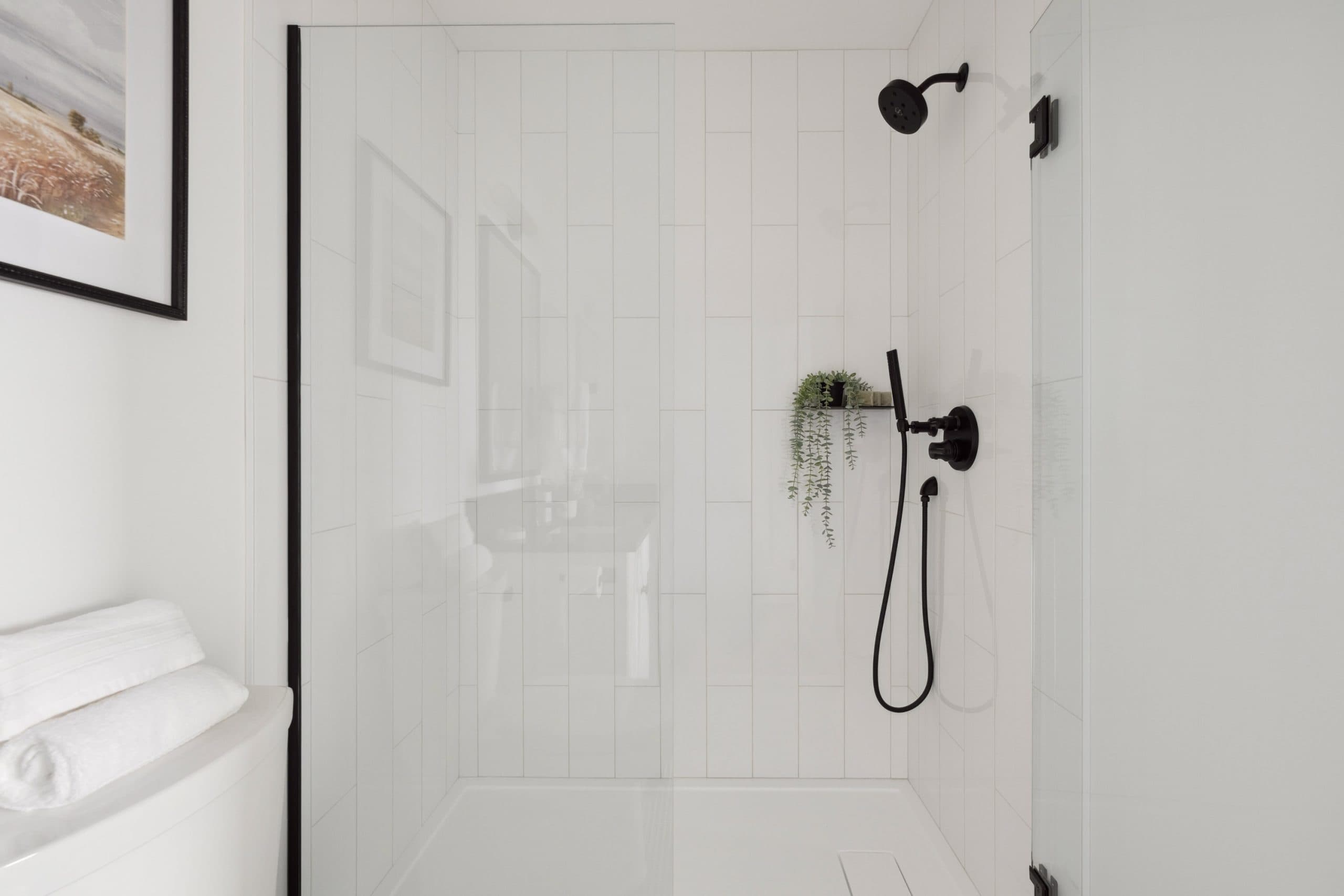 Water Tower Hill – A bright modern bathroom features a walk-in shower with clear glass doors and vertical white subway tiles. Matte black shower fixtures and hardware contrast against the monochromatic white walls, complemented by a small potted plant on a minimalist shelf.
