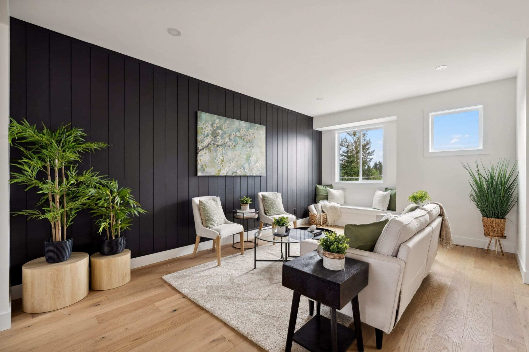 Water Tower Hill – A modern living room features a striking dark vertical wood slat accent wall adorned with a large canvas painting of white blossoms. The space is styled with light wood flooring, a cream sofa and armchairs, potted indoor greenery, and a cozy built-in window bench seat overlooking treetops under a clear sky.