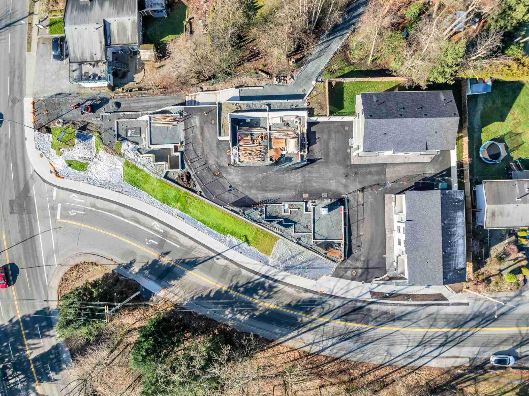 Water Tower Hill – A high-angle drone photograph shows a small residential development site under active construction, situated along a curving paved road. Several building foundations with concrete footings and framing materials sit adjacent to two completed or near-complete townhomes with dark shingled roofs.