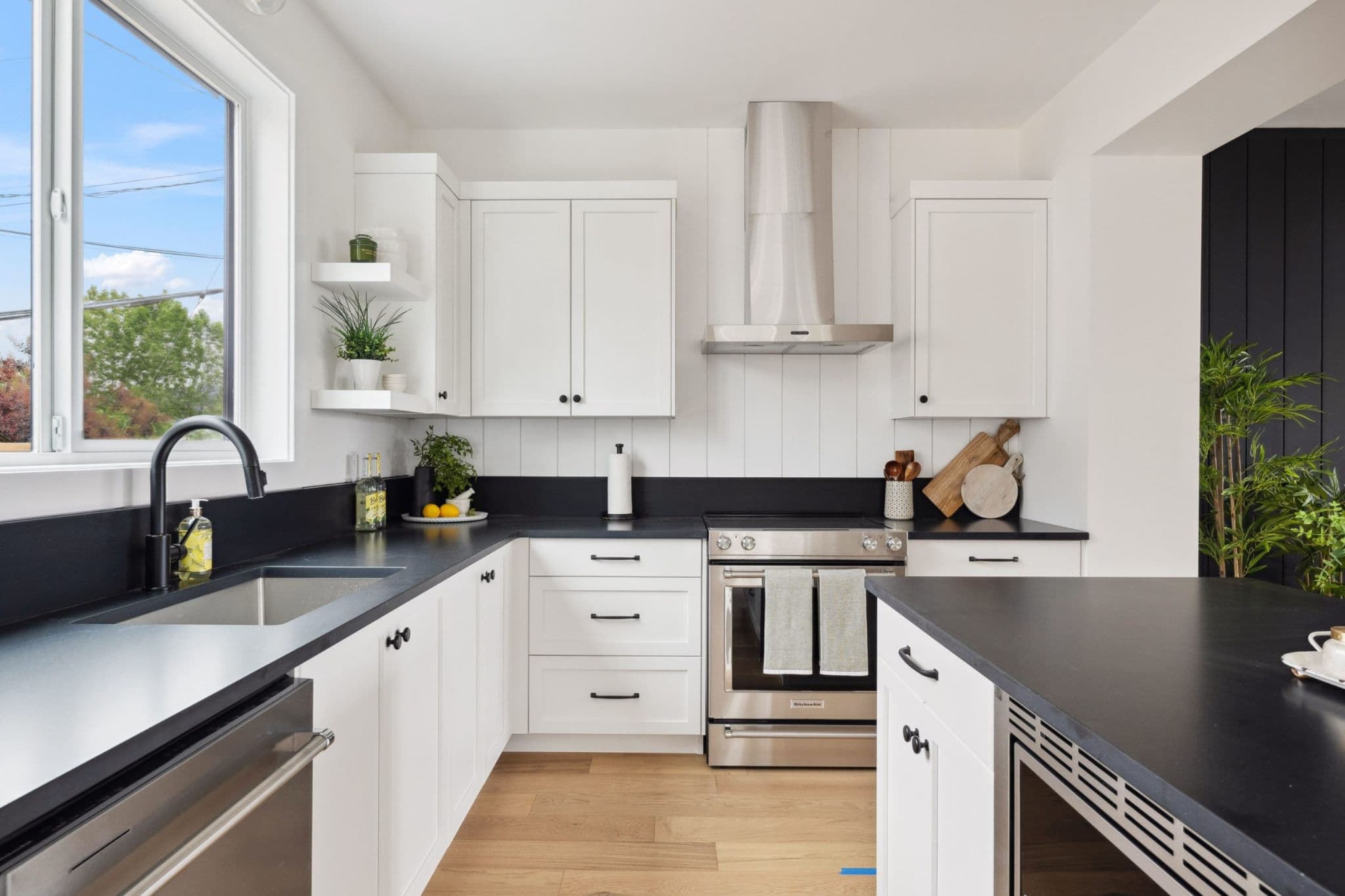 Water Tower Hill – A bright, modern kitchen features white shaker cabinetry paired with contrasting black countertops and a low black backsplash. Stainless steel appliances, including a slide-in range with a sleek chimney hood, are complemented by light wood floors and a window letting in natural daylight.