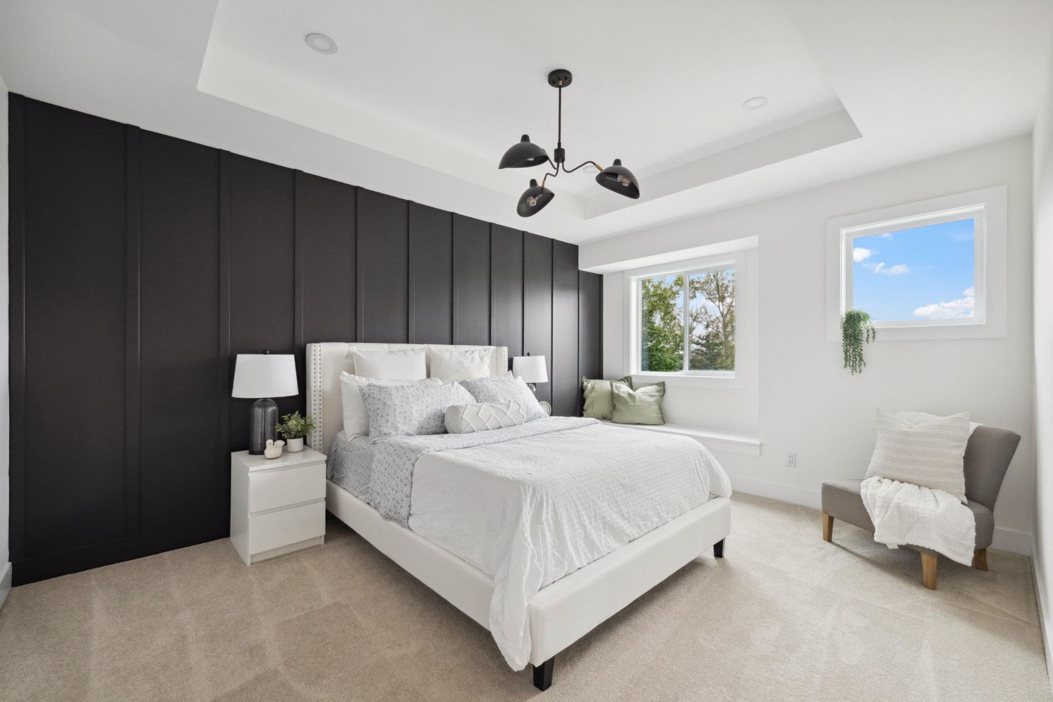 Water Tower Hill – A bright modern bedroom featuring a bed dressed in white linens centered against a dramatic black vertical board-and-batten accent wall. The room includes a tray ceiling with a black multi-arm light fixture, light neutral carpeting, a built-in window bench, and an armchair tucked in the corner.